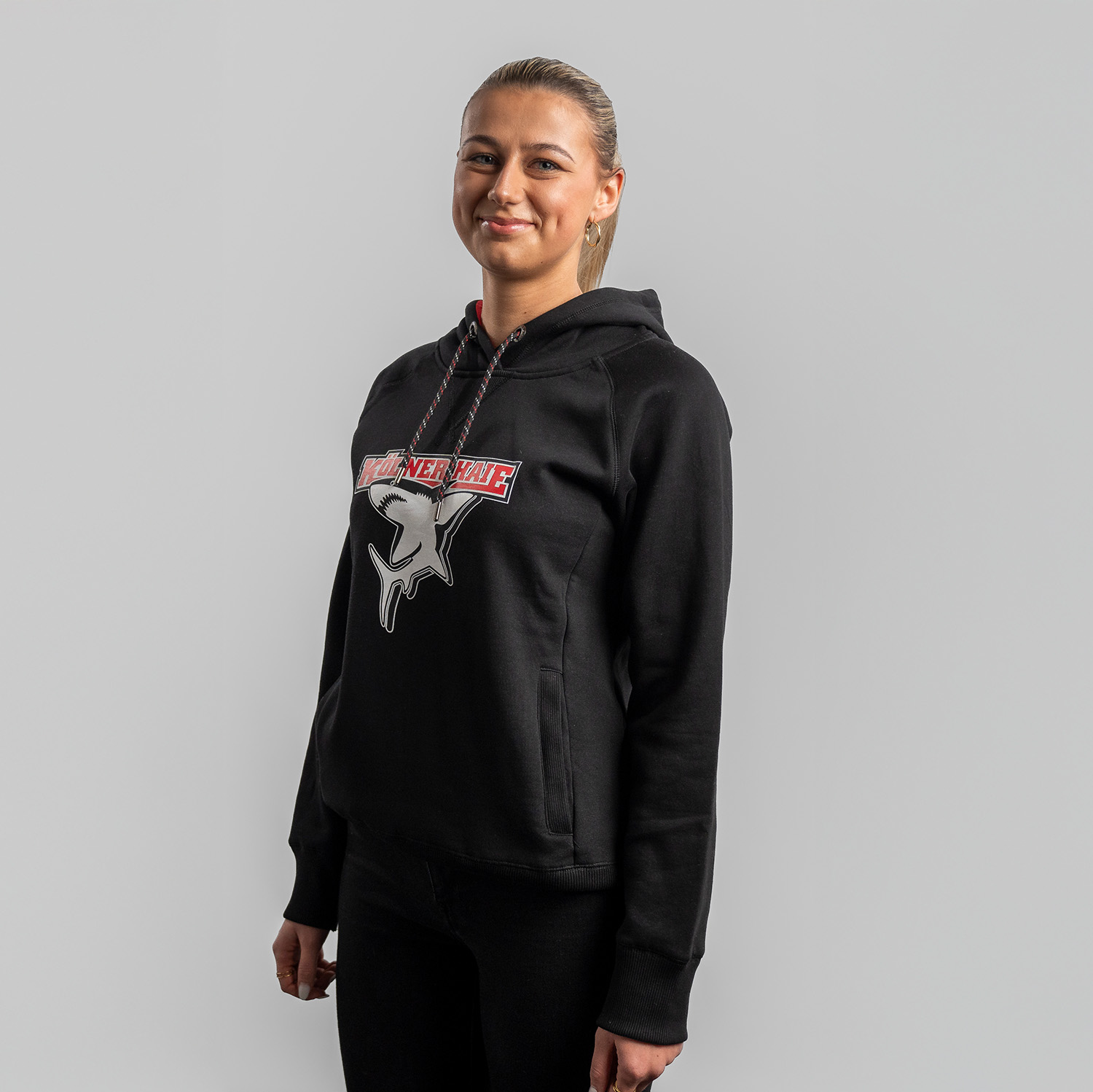 Schwarzer Damen Hoodie mit großem Frontprint vom Kölner Haie-Logo, getragen von Model, Seitansicht, linke Schulter vorne, vor hellem Hintergrund.