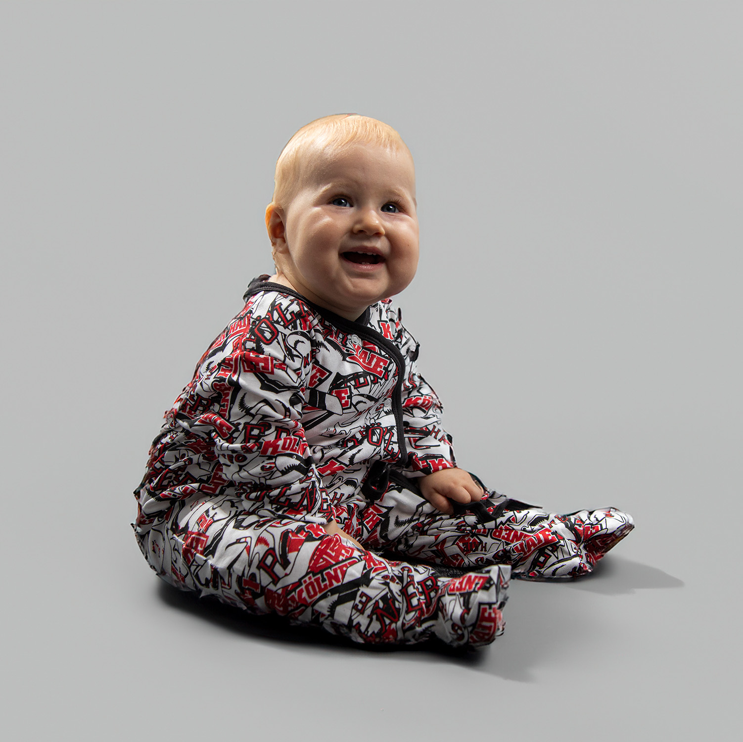 Baby Body EVOLUTION Schwarz-rot-weißer Kölner Haie-Body mit Allover-Print von verschiedenen Kölner Haie-Logos, getragen von Babymodel, sitzend vor hellem Hintergrund.