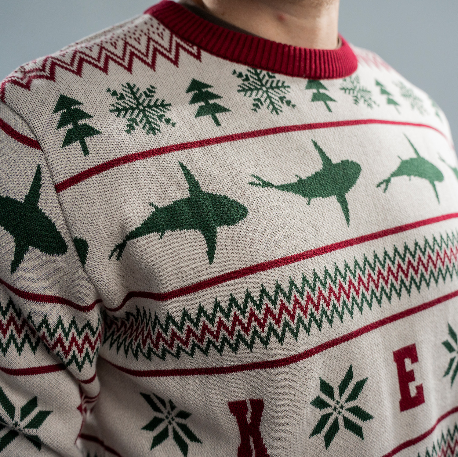 Detailansicht von weihnachtlich gestalteten Strickpullover in Beige mit roten und grünen Mustern. Der Pullover zeigt Reihen mit stilisierten Haien in grün, Schneeflocken, Tannenbäumen. Bündchen, Kragen und Saum sind in Dunkelrot abgesetzt.