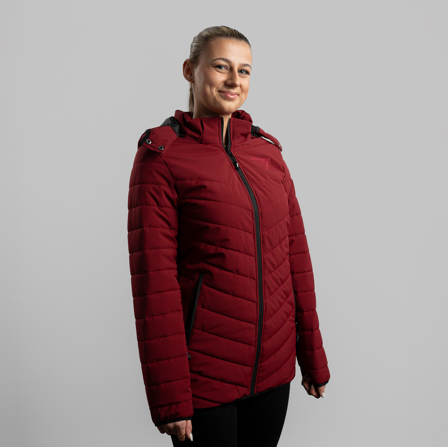 Bordeauxrote Damen Steppjacke mit abnehmbarer Kapuze, dezentes Ton-in-Ton Kölner Haie-Logo auf linker Brust (Herzseite) und zwei Eingrifftaschen, getragen von Model, Seitansicht 45 Grad, rechte Schulter vorne, vor hellem Hintergrund.