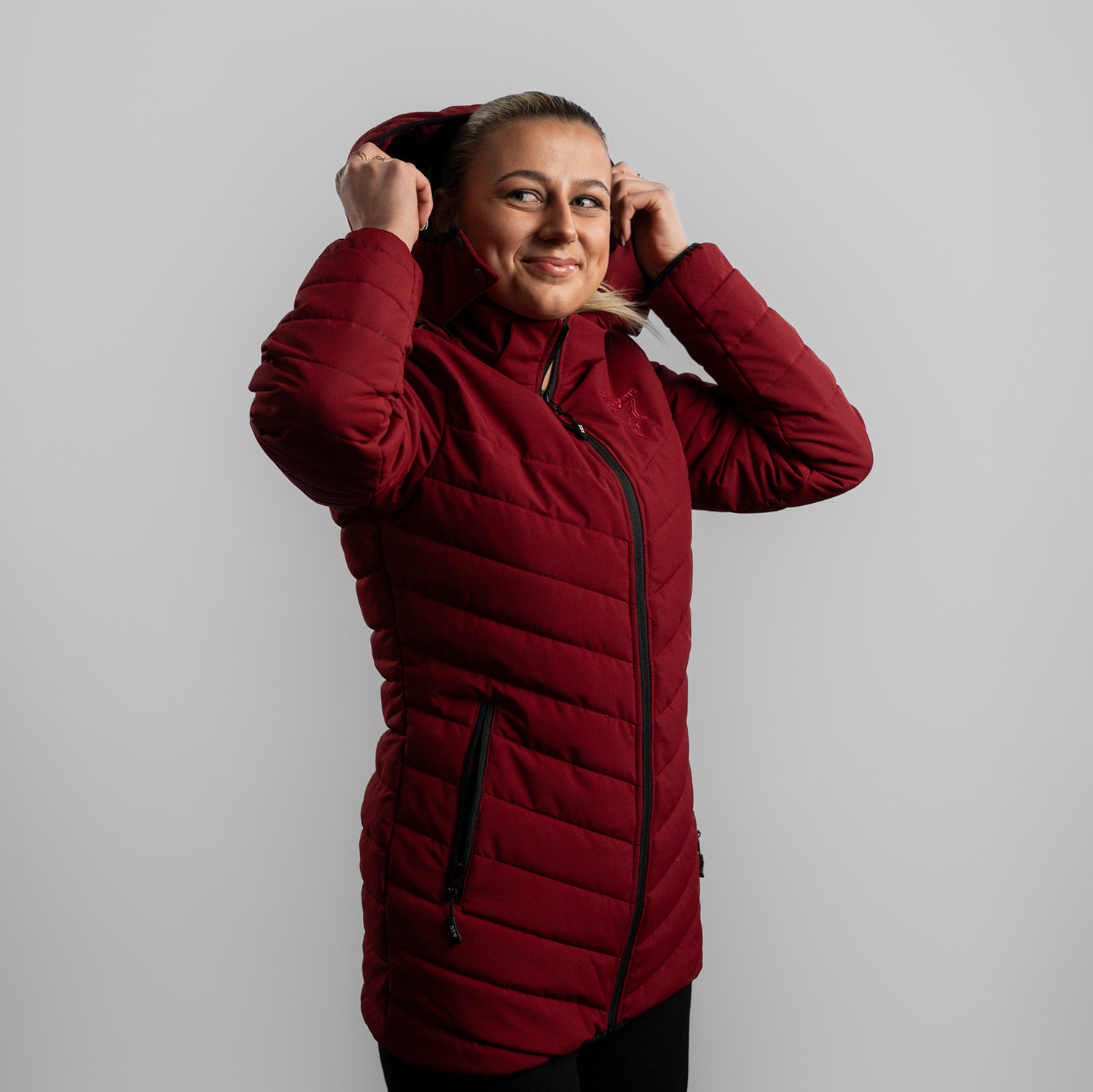 Bordeauxrote Damen Steppjacke mit abnehmbarer Kapuze, dezentes Ton-in-Ton Kölner Haie-Logo auf linker Brust (Herzseite) und zwei Eingrifftaschen, getragen von Model, Seitansicht 45 Grad, rechte Schulter vorne, Hände heben Kapuze über den Kopf, vor hellem