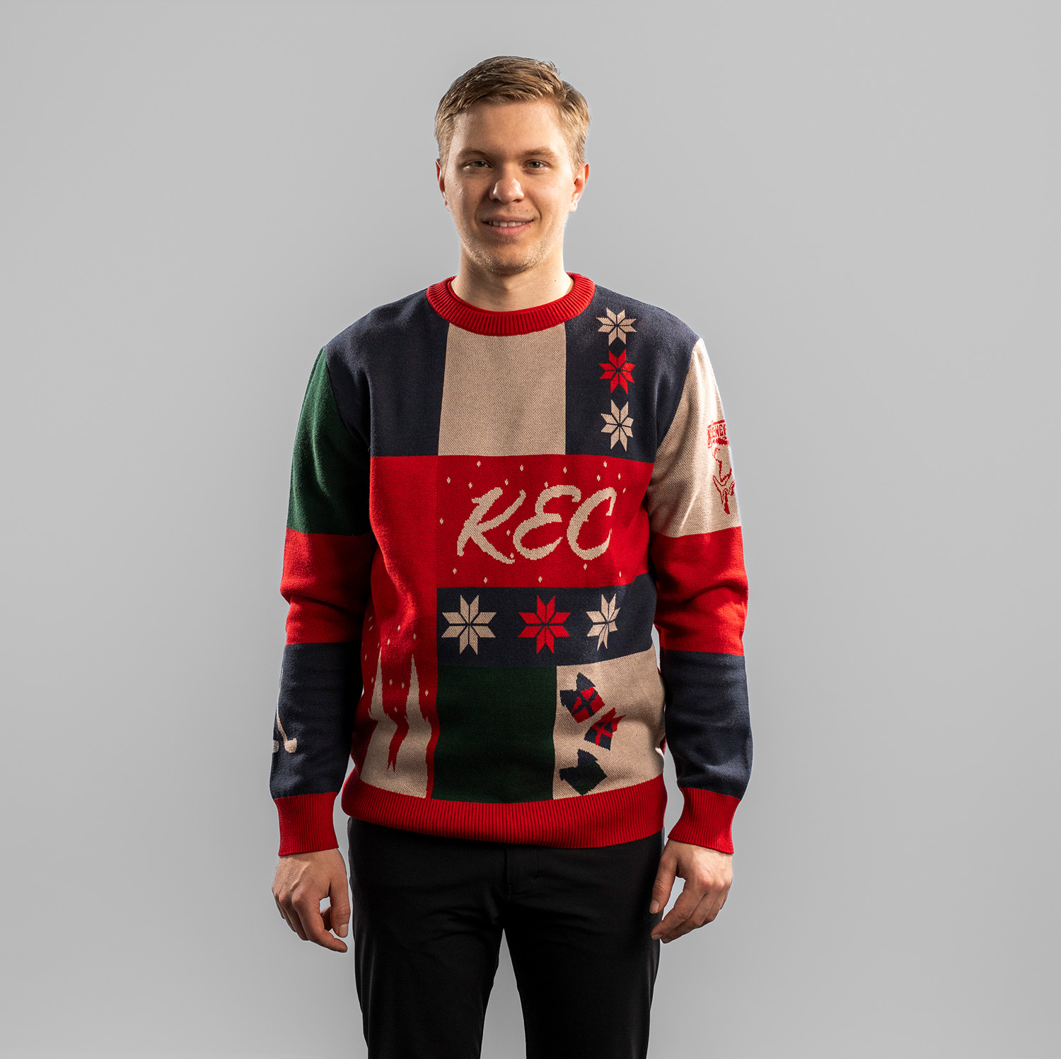 Tobias Ancicka trägt einen farbenfrohen Weihnachtspullover mit Patchwork-Design in Dunkelblau, Rot, Grün und Beige. Auf der Vorderseite steht der Schriftzug ‚KEC‘ in geschwungener Schrift, umgeben von Schneeflocken, Geschenken, dem Kölner Dom und stilisie