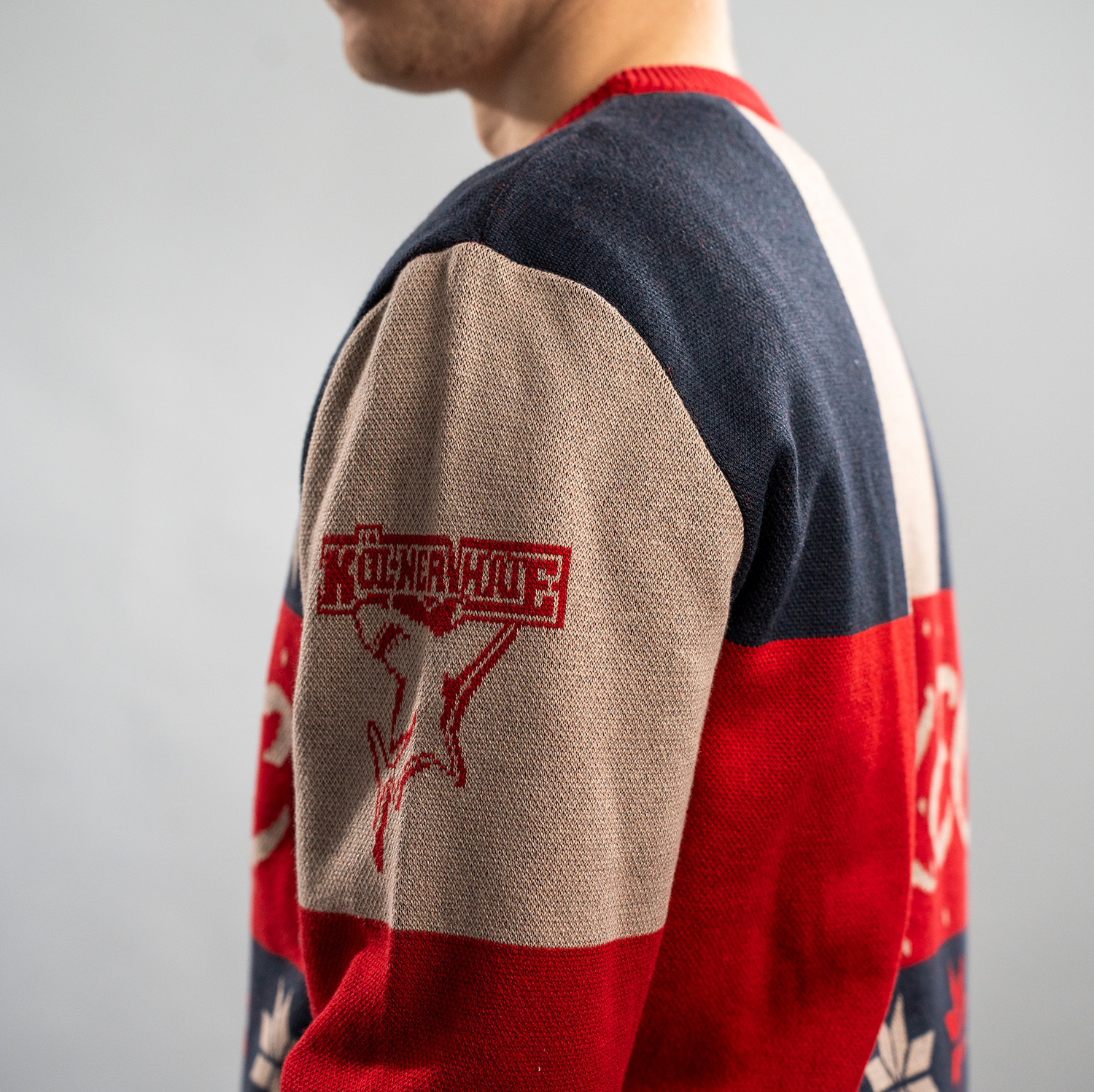 Detailansicht des roten Kölner Haie Logos auf dem linken Ärmel des farbenfrohen Weihnachtspullover mit Patchwork-Design in Dunkelblau, Rot, Grün und Beige.