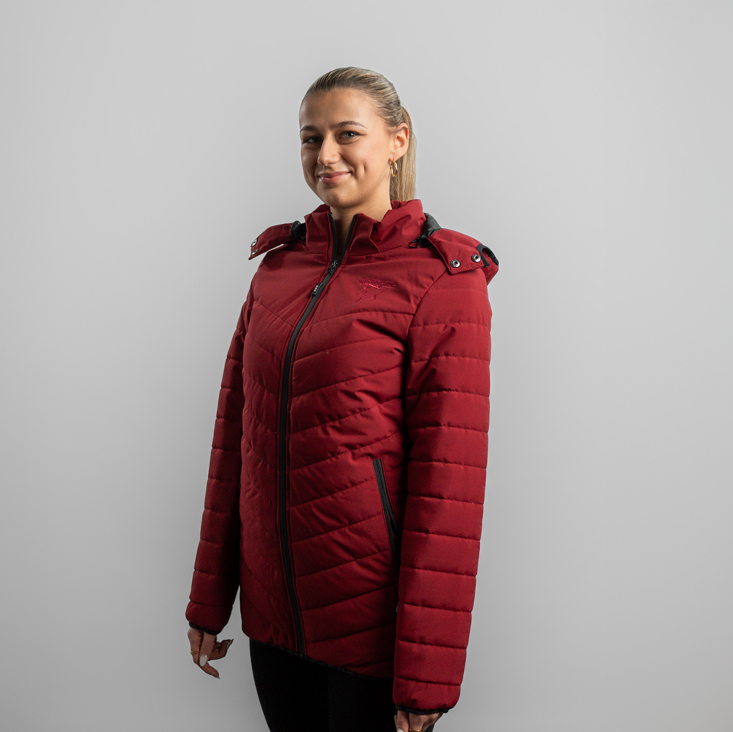 Bordeauxrote Damen Steppjacke mit abnehmbarer Kapuze, dezentes Ton-in-Ton Kölner Haie-Logo auf linker Brust (Herzseite) und zwei Eingrifftaschen, getragen von Model, Seitansicht 45 Grad, linke Schulter vorne, vor hellem Hintergrund.