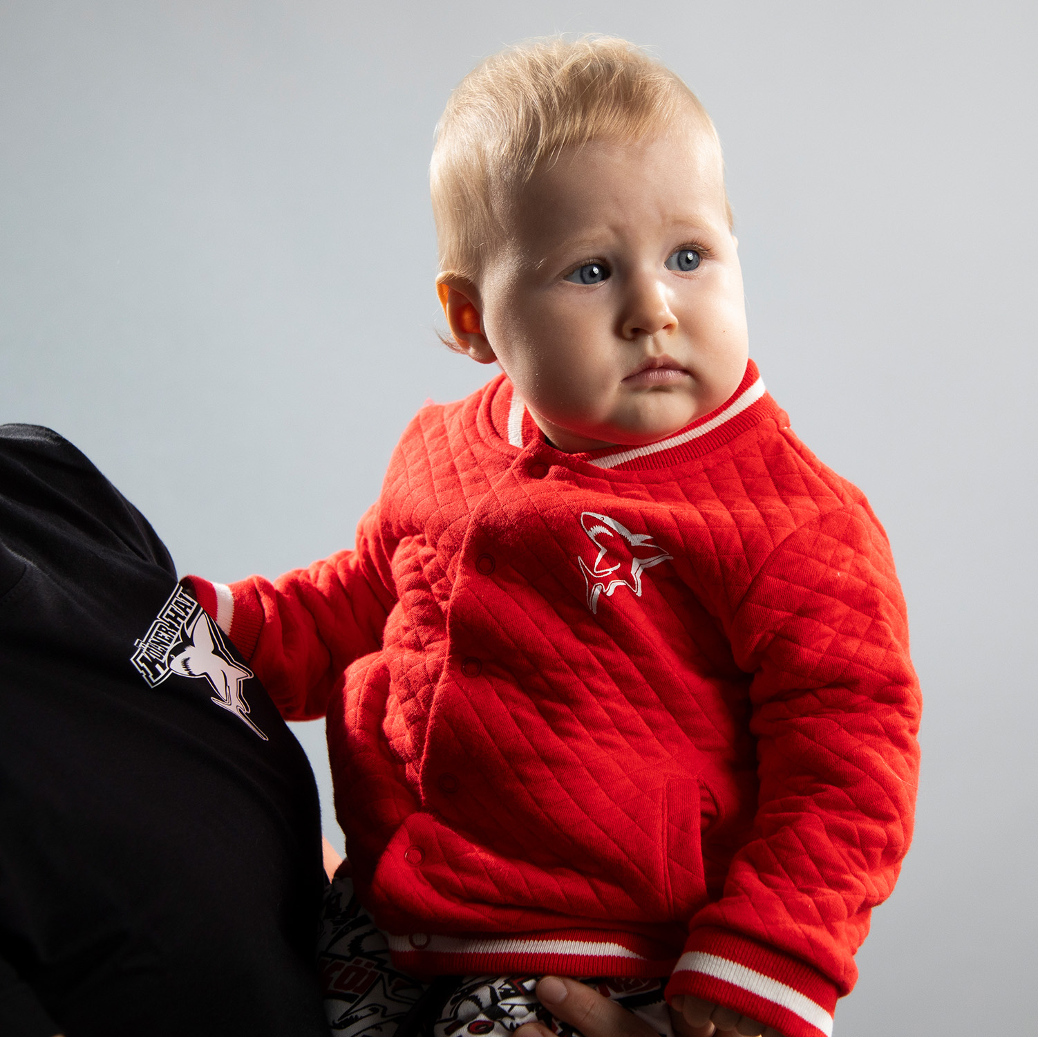 Rote Baby Jacke mit Knopfleiste und weißem Hai-Logo auf der linken Brustseite und weißen Highlights an Kragen und Säumen, getragen von Babymodel, sitzend in Elternarm vor hellem Hintergrund.