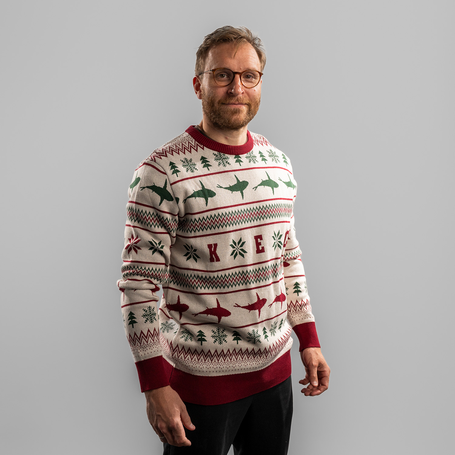 Frederik Storm trägt einen weihnachtlich gestalteten Strickpullover in Beige mit roten und grünen Mustern. Der Pullover zeigt Reihen mit stilisierten Haien, Schneeflocken, Tannenbäumen und den Buchstaben ‚K E C‘ auf Brusthöhe. Bündchen, Kragen und Saum si