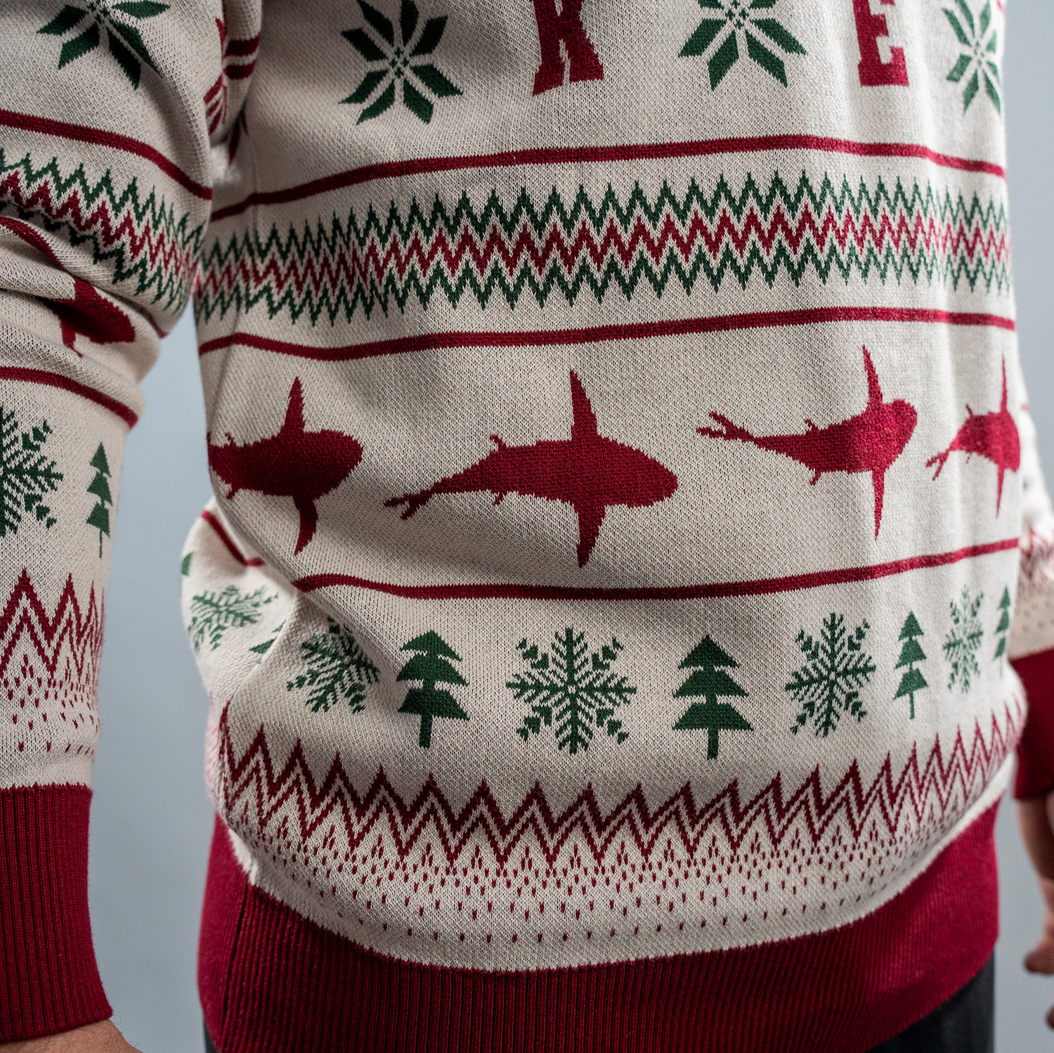 Detailansicht von weihnachtlich gestalteten Strickpullover in Beige mit roten und grünen Mustern. Der Pullover zeigt Reihen mit stilisierten Haien in dunkelrot, Schneeflocken, Tannenbäumen. Bündchen, Kragen und Saum sind in Dunkelrot abgesetzt.