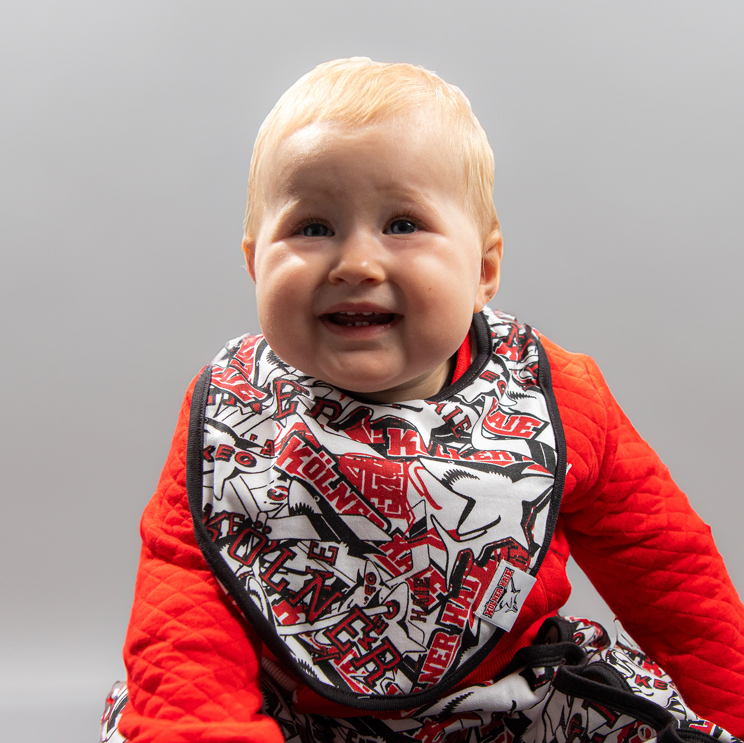 Lätzchen HAIE Schwarz-rot-weißes Kölner Haie-Lätzchen mit Allover-Print von verschiedenen Kölner Haie-Logos, getragen von Babymodel, sitzend vor hellem Hintergrund.