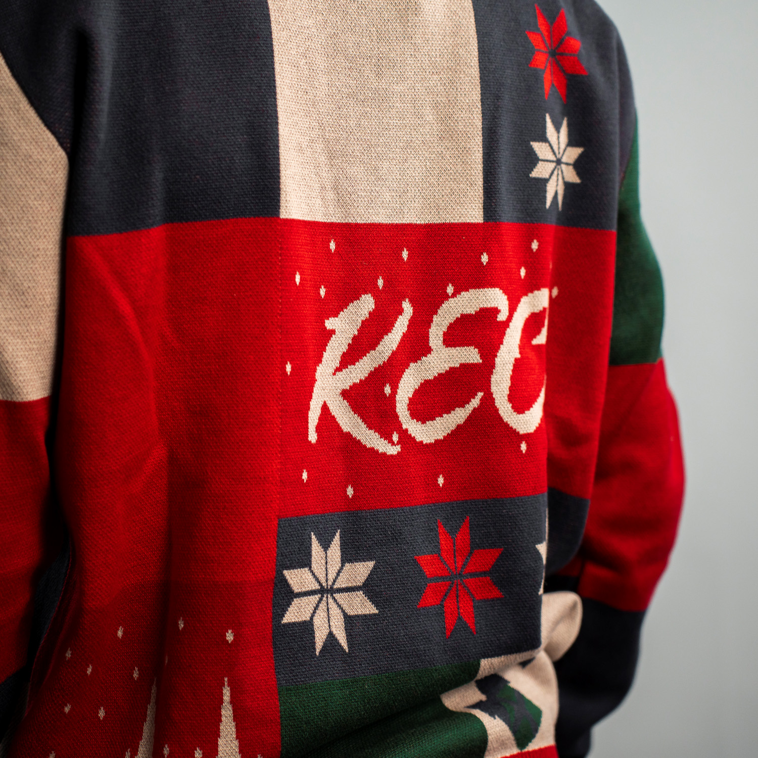Detailansicht "KEC"-Schriftzug in geschwungener Schrift auf dem farbenfrohen Weihnachtspullover mit Patchwork-Design in Dunkelblau, Rot, Grün und Beige.  