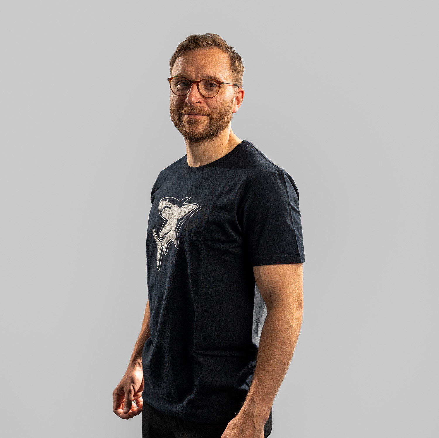 Navy-blaues T-Shirt der Kölner Haie mit großem Frontprint des Hais aus dem Kölner Haie-Logo. Getragen von Frederik Storm, Seitansicht, linke Schulter vorne, vor hellem Hintergrund.