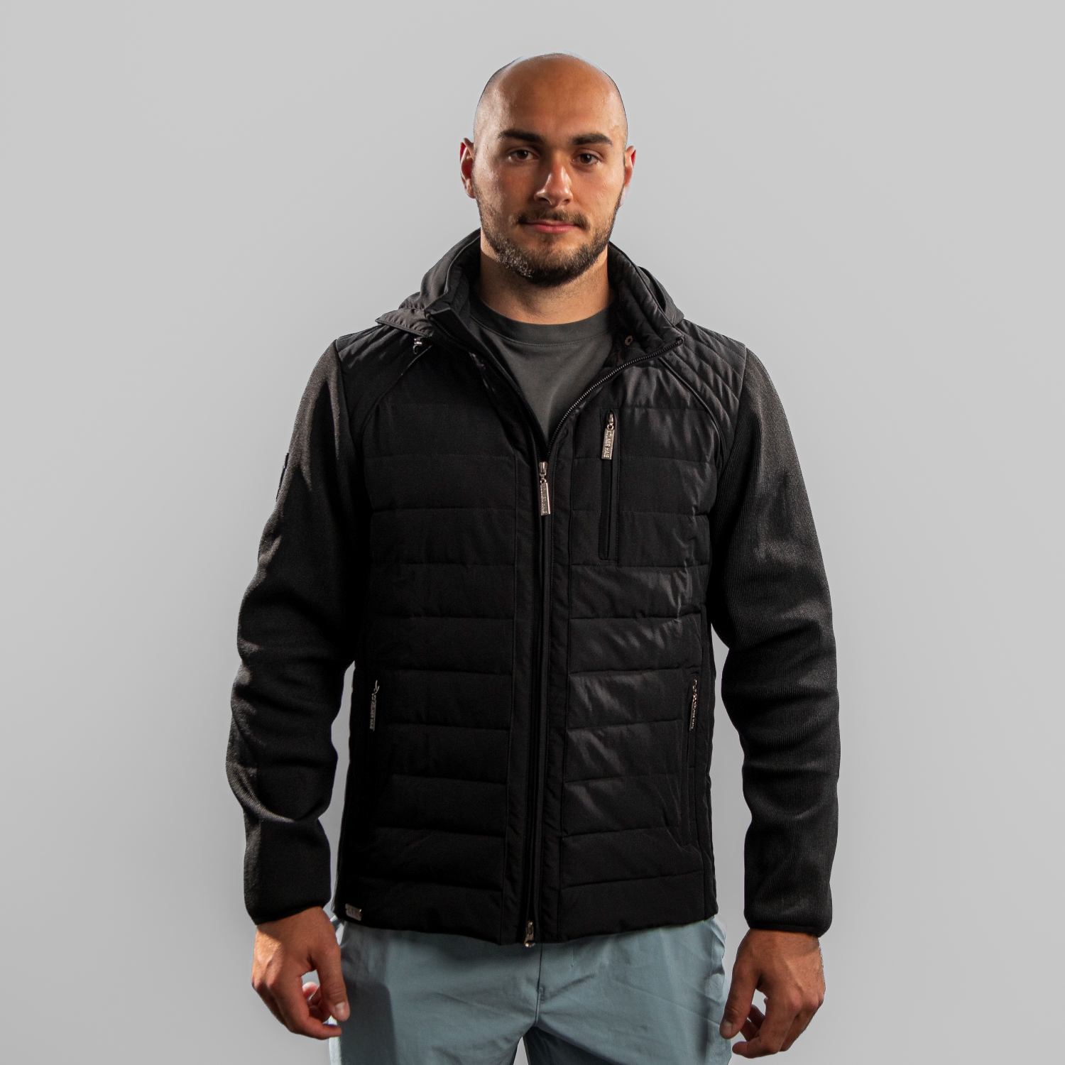 Schwarze Hybrid-Übergangsjacke mit Ton-in-Ton Patch vom Kölner Haie-Logo auf rechter Schulter, getragen von Maxi Glötzl. Der Body ist in einem Padded-Look, die Ärmel abgesetzt aus anderem Material. Frontansicht, vor hellem Hintergrund.