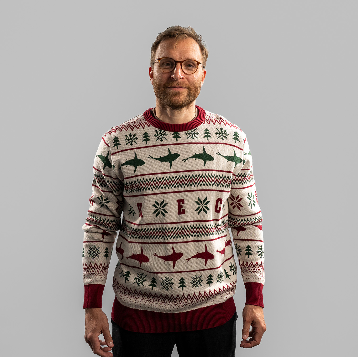 Frederik Storm trägt einen weihnachtlich gestalteten Strickpullover in Beige mit roten und grünen Mustern. Der Pullover zeigt Reihen mit stilisierten Haien, Schneeflocken, Tannenbäumen und den Buchstaben ‚K E C‘ auf Brusthöhe. Bündchen, Kragen und Saum si
