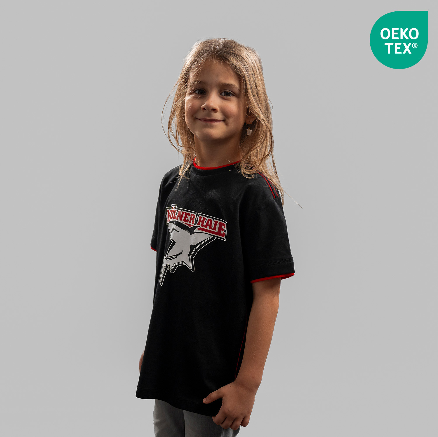 T-Shirt BASIC kids Schwarzes Kinder T-Shirt mit großem Frontprint vom Kölner Haie-Logo, getragen von Kindermodel, Seitansicht, linke Schulter vorne, vor hellem Hintergrund. Der Artikel ist OEKO-TEX® MADE IN GREEN zertifiziert.