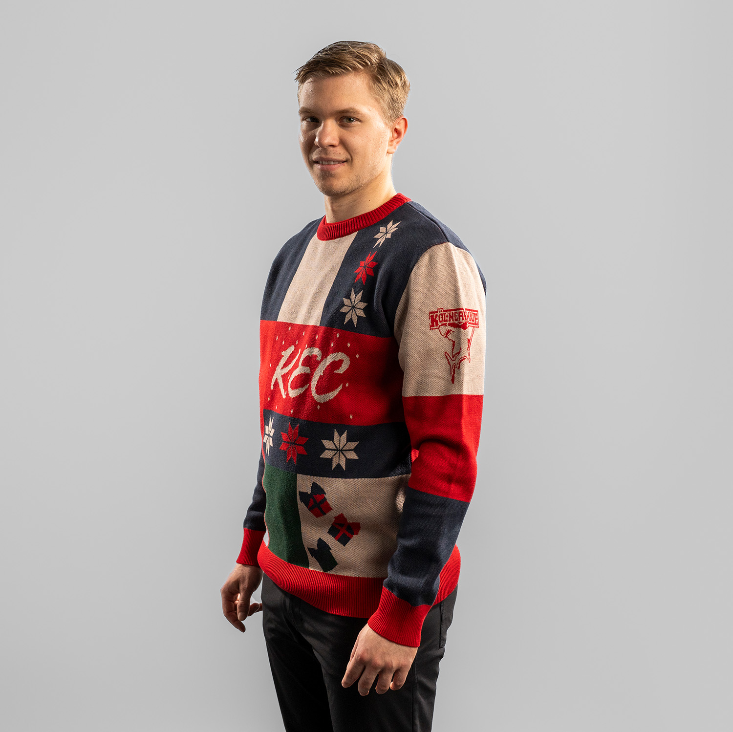 Tobias Ancicka trägt einen farbenfrohen Weihnachtspullover mit Patchwork-Design in Dunkelblau, Rot, Grün und Beige. Auf der Vorderseite steht der Schriftzug ‚KEC‘ in geschwungener Schrift, umgeben von Schneeflocken, Geschenken, Strümpfen und stilisierten 