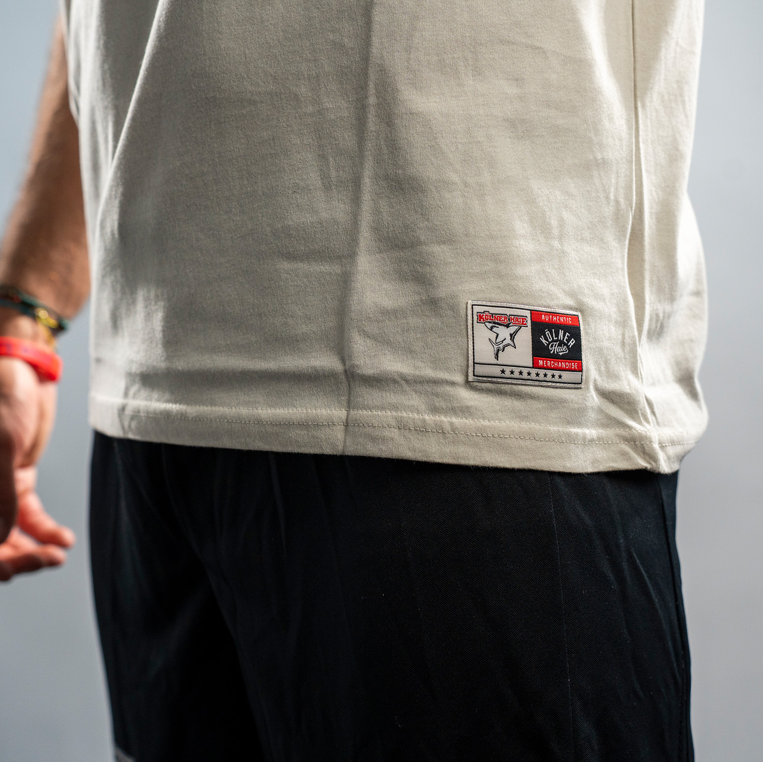 Detailansicht zweigeteiltes Web-Label am unteren linken Bündchen. Linke Seite Kölner Haie-Logo, rechte Seite von oben nach untenstehend: Authentic Kölner Haie Merchandising. Darunter sind 8 Sterne. 