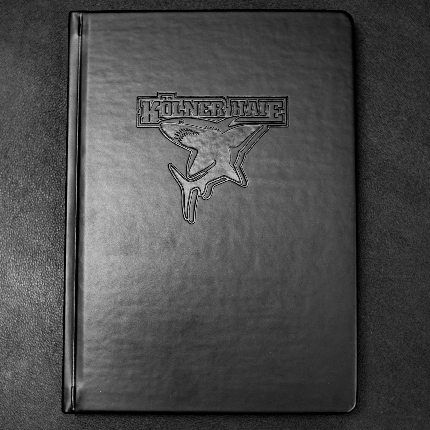 Schwarzes Notizbuch mit Ton-in-Ton geprägtem Kölner Haie-Logo auf dem Cover, liegend auf dunklem Untergrund, Draufansicht