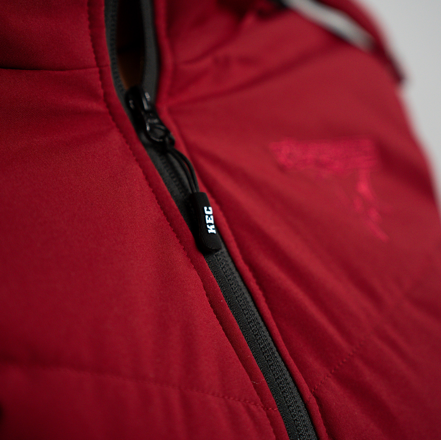Detailansicht schwarzer Zipper mit KEC Schriftzug der bordeauxroter Jacke.