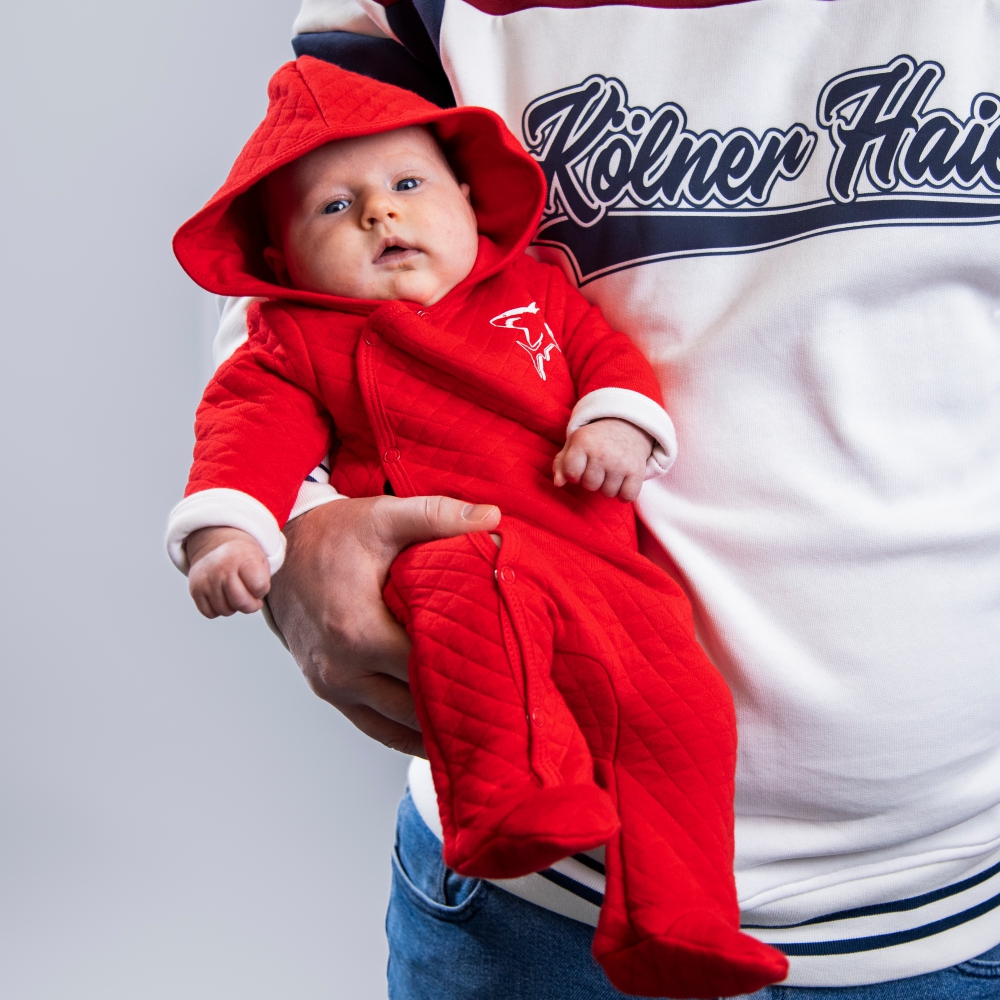 Overall ROOKIE kids Roter Baby Overall mit Kapuze, Füßen und weißem Hai-Logo auf der linken Brustseite und weißen umgeklappten Ärmelsaum, getragen von Babymodel, liegend in Elternarm vor hellem Hintergrund.