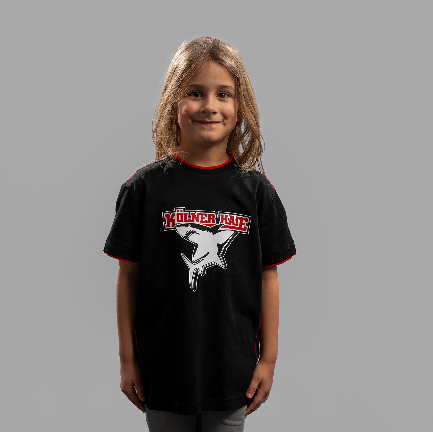 Schwarzes Kinder T-Shirt mit großem Frontprint vom Kölner Haie-Logo, getragen von Kindermodel, Frontansicht, vor hellem Hintergrund.