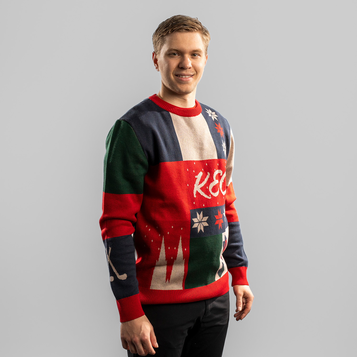 Ugly Sweater RUDSLÄTT Tobias Ancicka trägt einen farbenfrohen Weihnachtspullover mit Patchwork-Design in Dunkelblau, Rot, Grün und Beige. Auf der Vorderseite steht der Schriftzug ‚KEC‘ in geschwungener Schrift, umgeben von Schneeflocken, Geschenken, Strümpfen und stilisierten