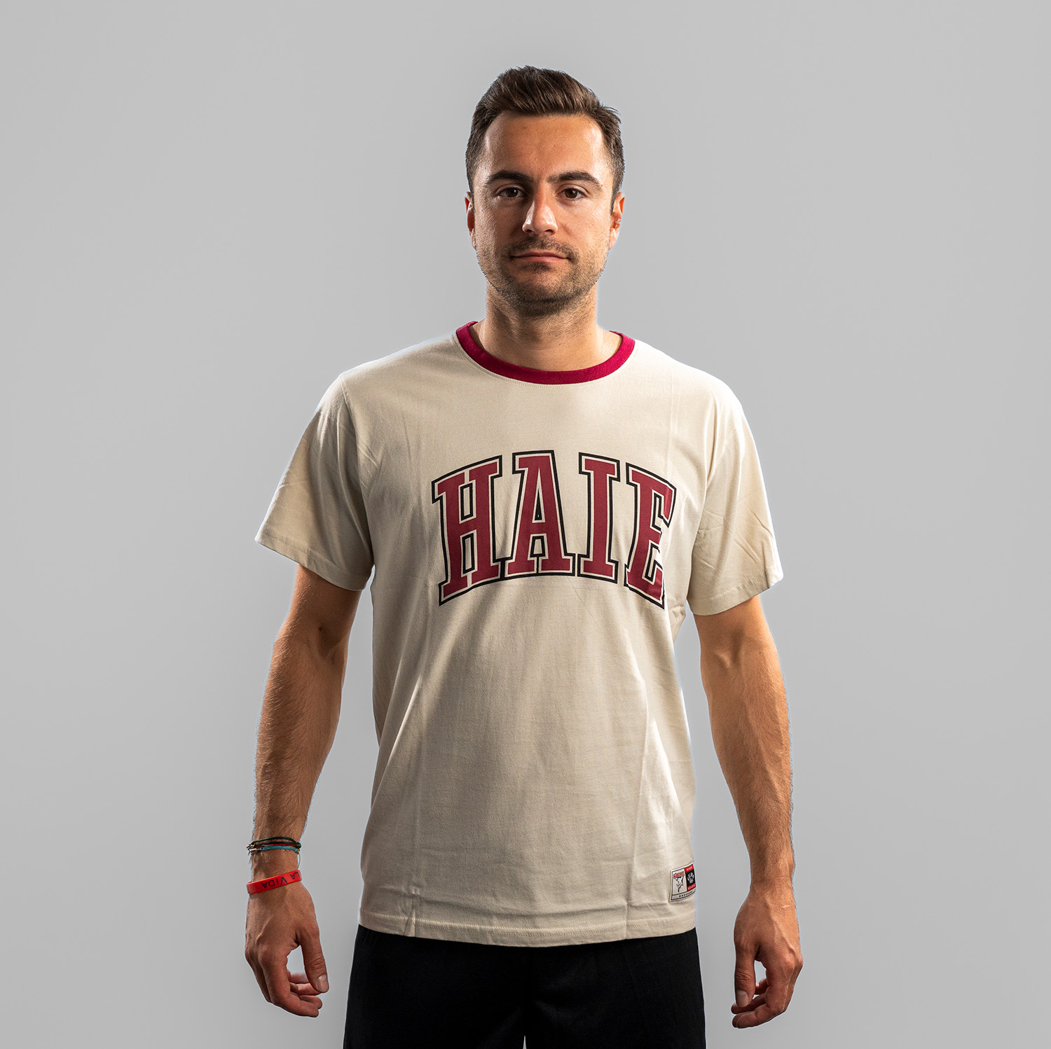 Beiges T-Shirt der Kölner Haie mit großem Frontprint des Schriftzuges „Haie“ in bordeauxrot und bordeauxrot abgesetzten Halsloch, getragen von Maxi Kammerer, Frontansicht, vor hellem Hintergrund.