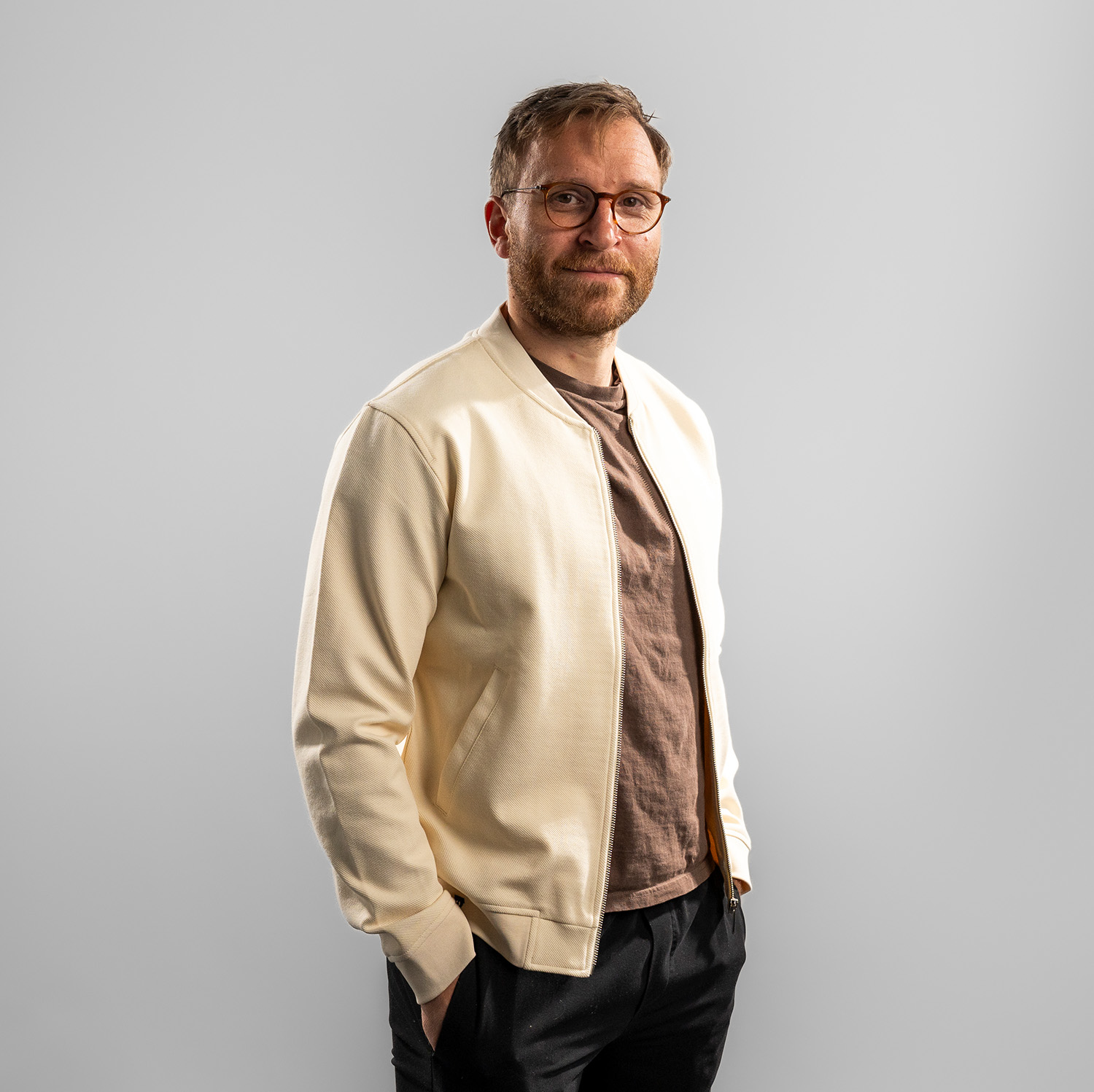 Jacke DANIEL Beigefarbene Collegejacke mit Reißverschluss und dezentem, tonal gesticktem Haie-Logo auf der linken Brustseite. Getragen von Frederik Storm, Frontansicht mit leichter Schultereindrehung, rechte Schulter vorne, vor hellem Hintergrund.