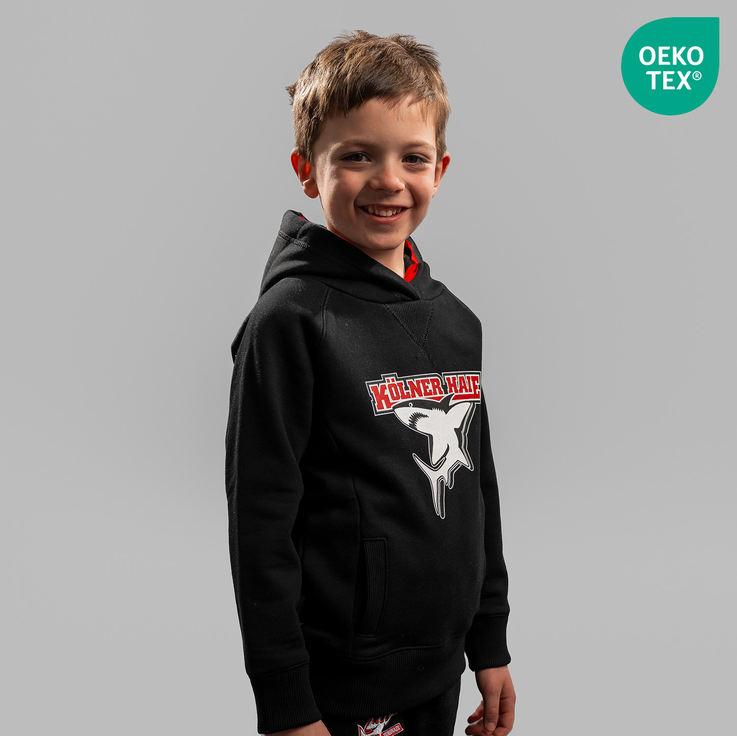 Hoodie BASIC kids Schwarzer Kinder Hoodie mit großem Frontprint vom Kölner Haie-Logo, getragen von Kindermodel, Seitansicht, rechte Schulter vorne, vor hellem Hintergrund. Der Artikel ist OEKO-TEX® MADE IN GREEN zertifiziert.