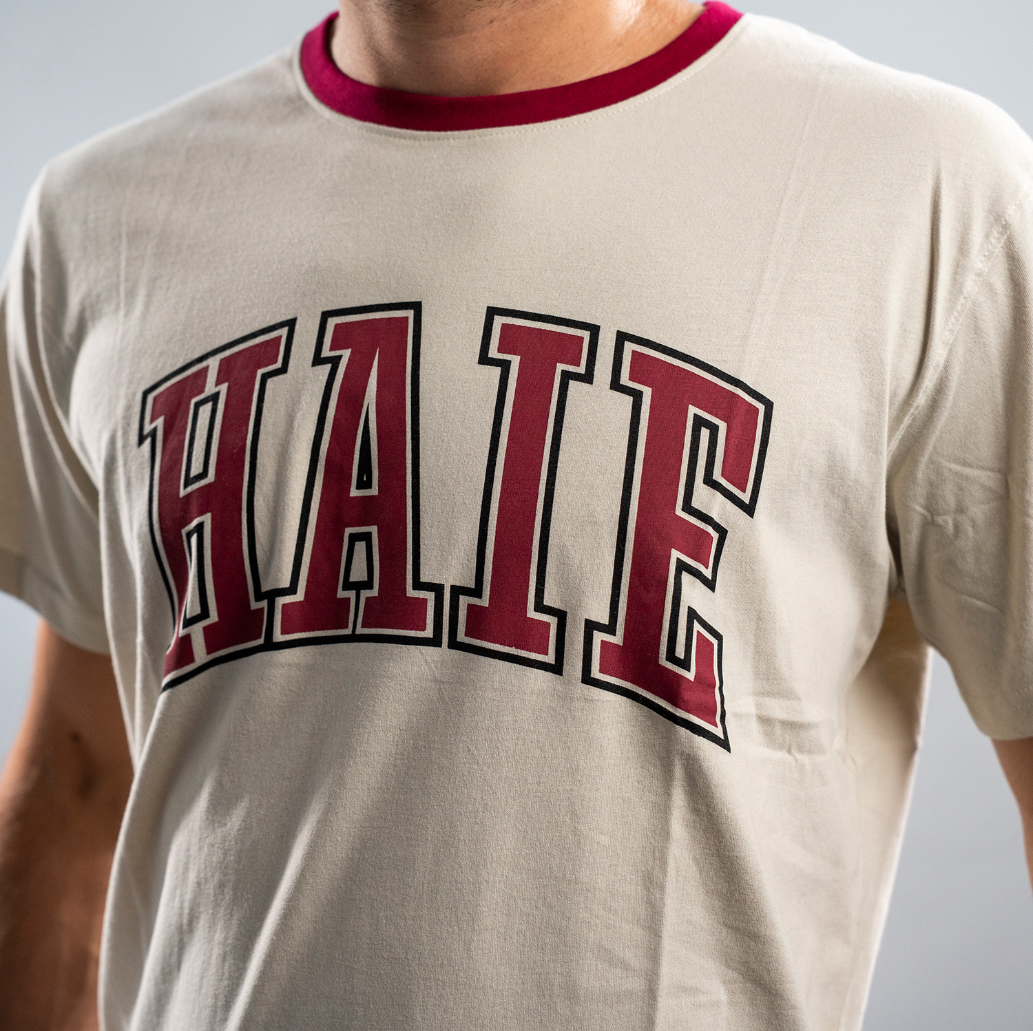 Detailansicht Druck vom „Haie“ auf dem beigen T-Shirt. Die Buchstaben sind bordeauxrot mit einer beigen und einer schwarzen Umrandung.