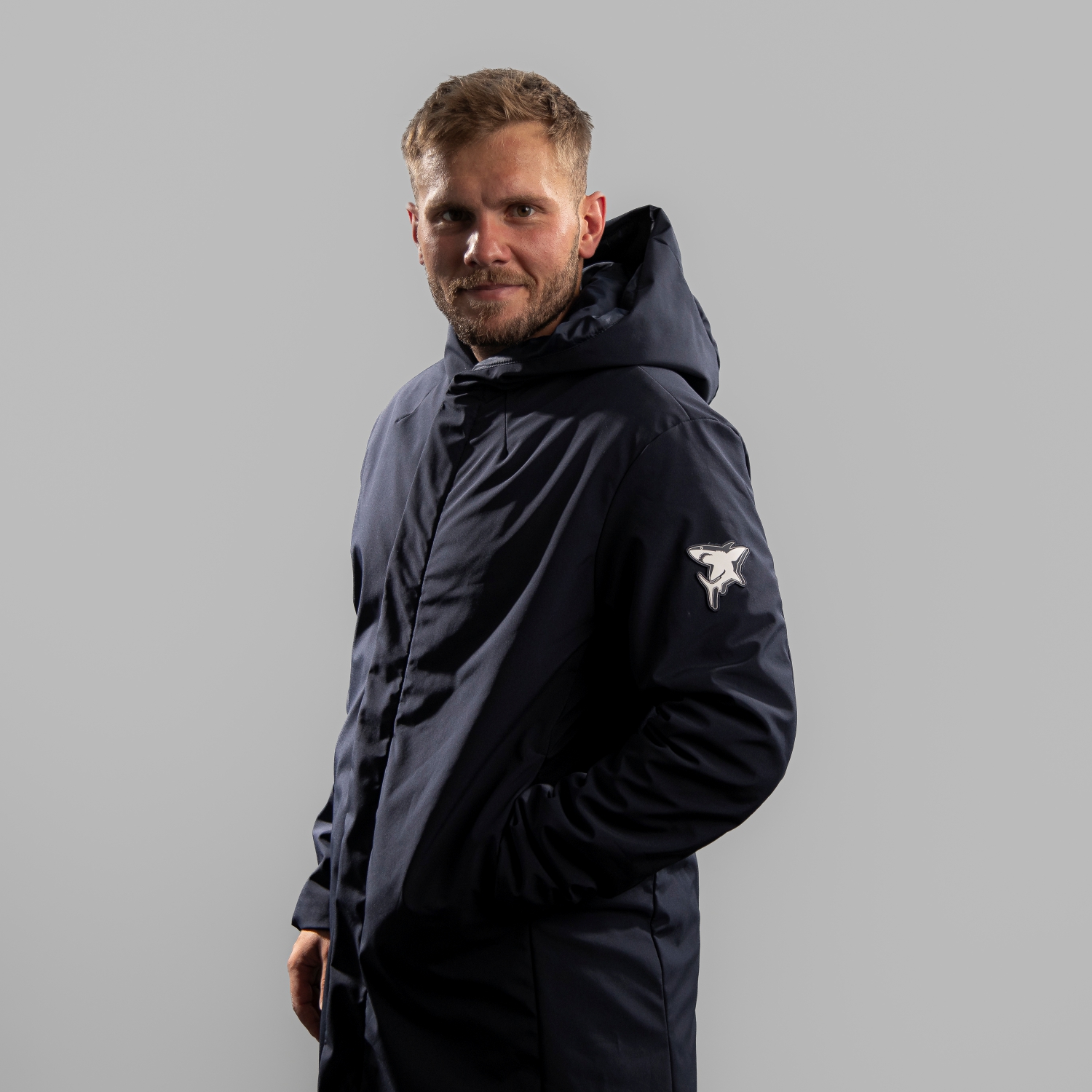 Parka SHARK Dunkelblauer schlichter Parka mit Silikon-Patch des Hai aus dem Kölner Haie-Logo auf dem linken Ärmel, getragen von Parker Tuomie, Seitansicht linke Schulter vorne, vor hellem Hintergrund.