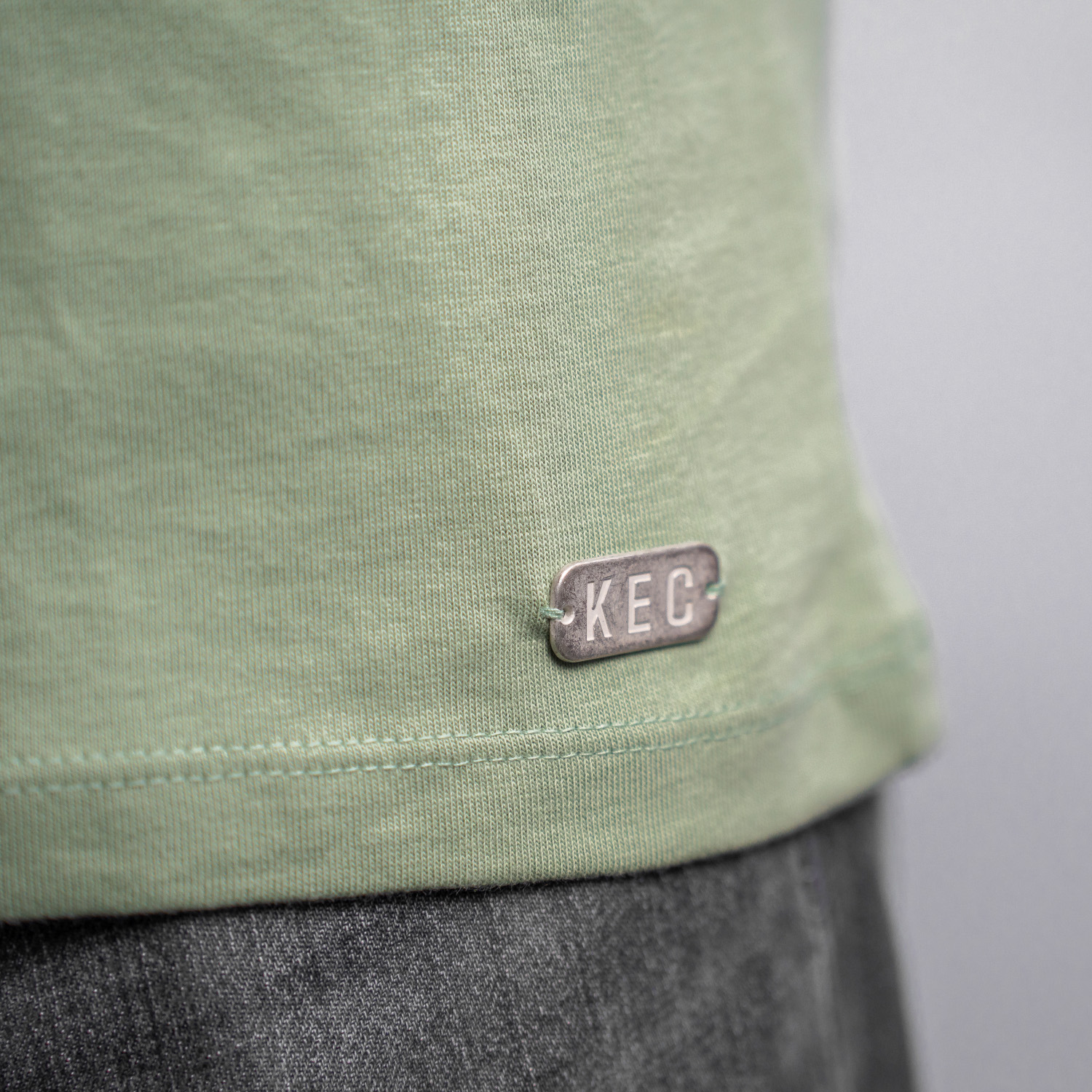 Detailansicht Metallplättchen am unteren linken Rand des grünen T-Shirts angenäht. Die Buchstaben KEC sind eingeprägt.