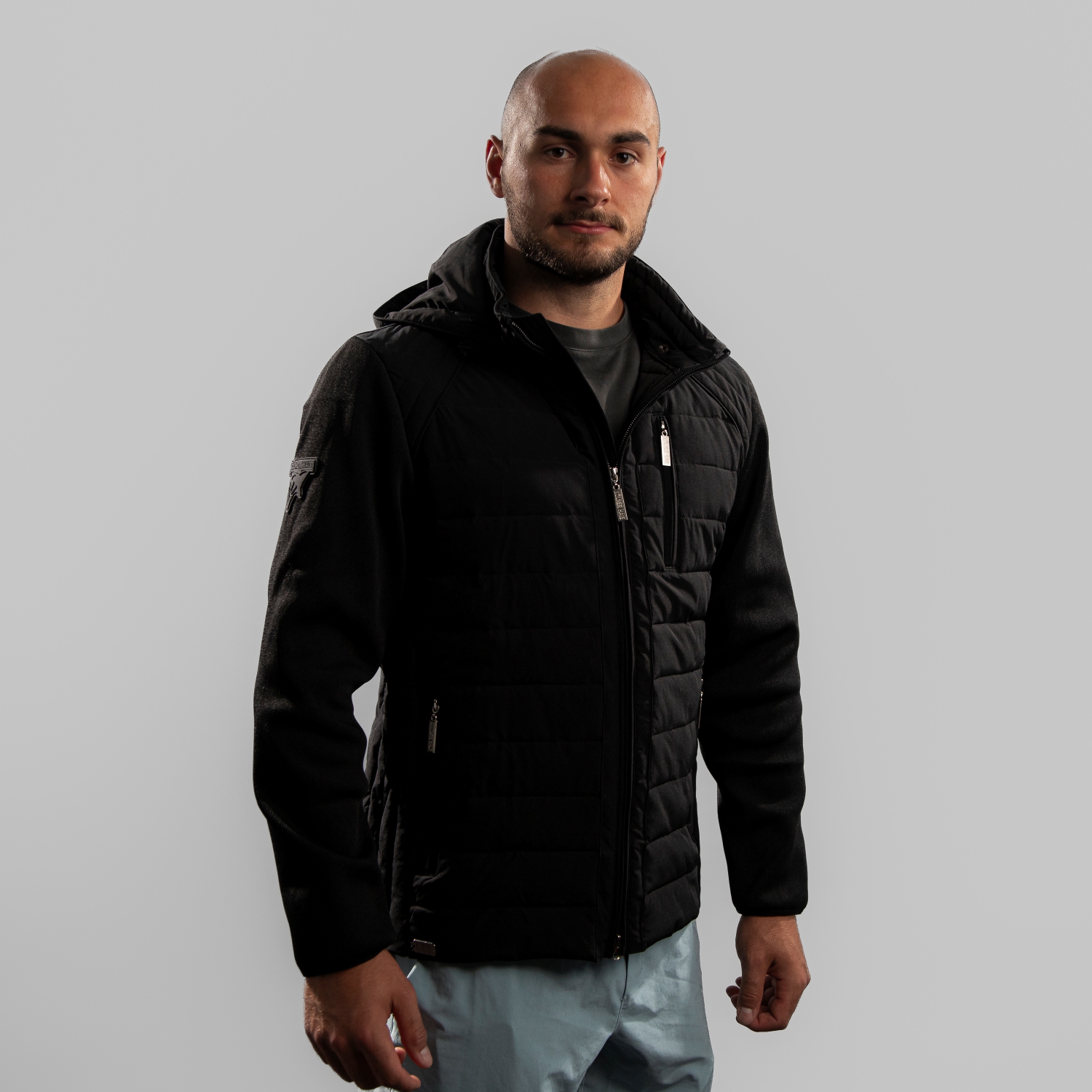 Jacke STYLE Schwarze Hybrid-Übergangsjacke mit Ton-in-Ton Patch vom Kölner Haie-Logo auf rechter Schulter, getragen von Maxi Glötzl. Der Body ist in einem Padded-Look, die Ärmel abgesetzt aus anderem Material. Seitansicht, rechte Schulter vorne, vor hellem Hintergrun