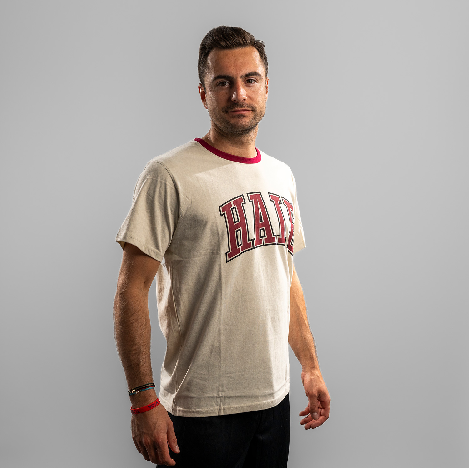 Beiges T-Shirt der Kölner Haie mit großem Frontprint des Schriftzuges „Haie“ in bordeauxrot und bordeauxrot abgesetzten Halsloch, getragen von Maxi Kammerer, Seitansicht, rechte Schulter vorne, vor hellem Hintergrund.