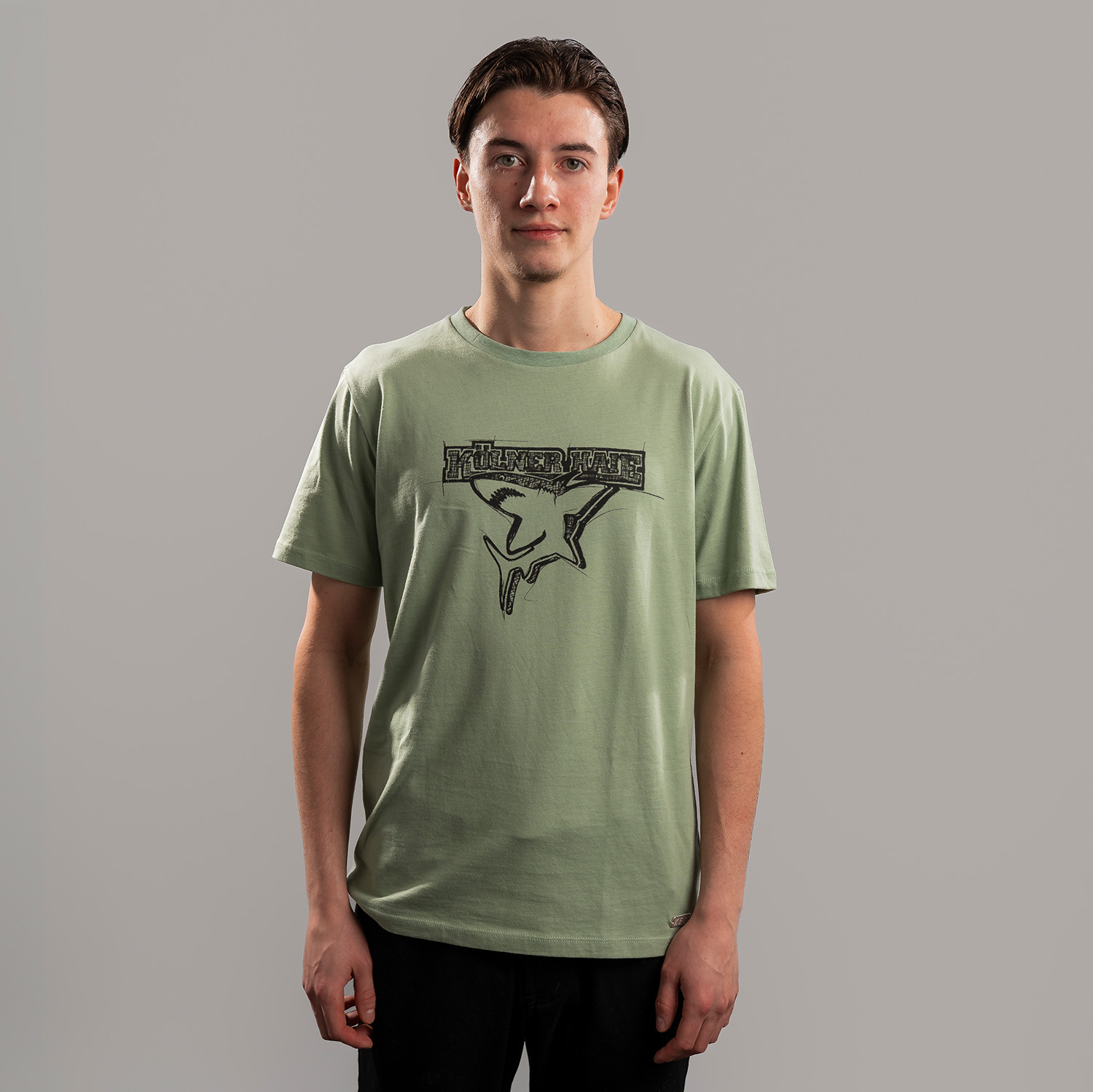 Grünes T-Shirt der Kölner Haie mit großem Frontprint des Kölner Haie-Logos, getragen von Kevin Niedenz, Frontansicht, vor hellem Hintergrund.