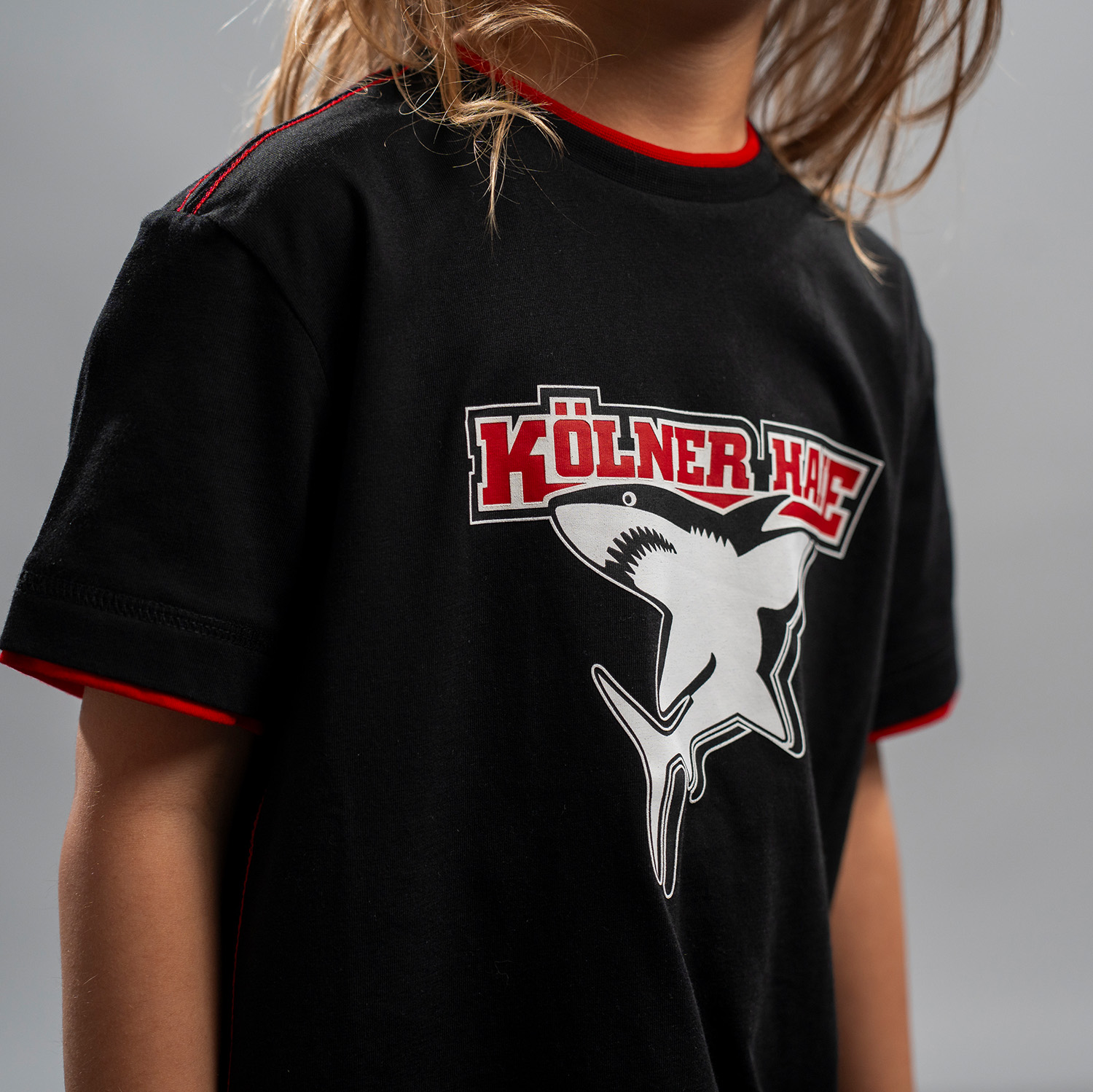 Detailansicht 3D-Druck vom Kölner Haie-Logo auf schwarzem Kinder T-Shirt. Am Halsloch und an den Ärmelbündchen ist roter Stoff eingesetzt. Die Nähte auf den Schultern und an den Seiten sind rot.