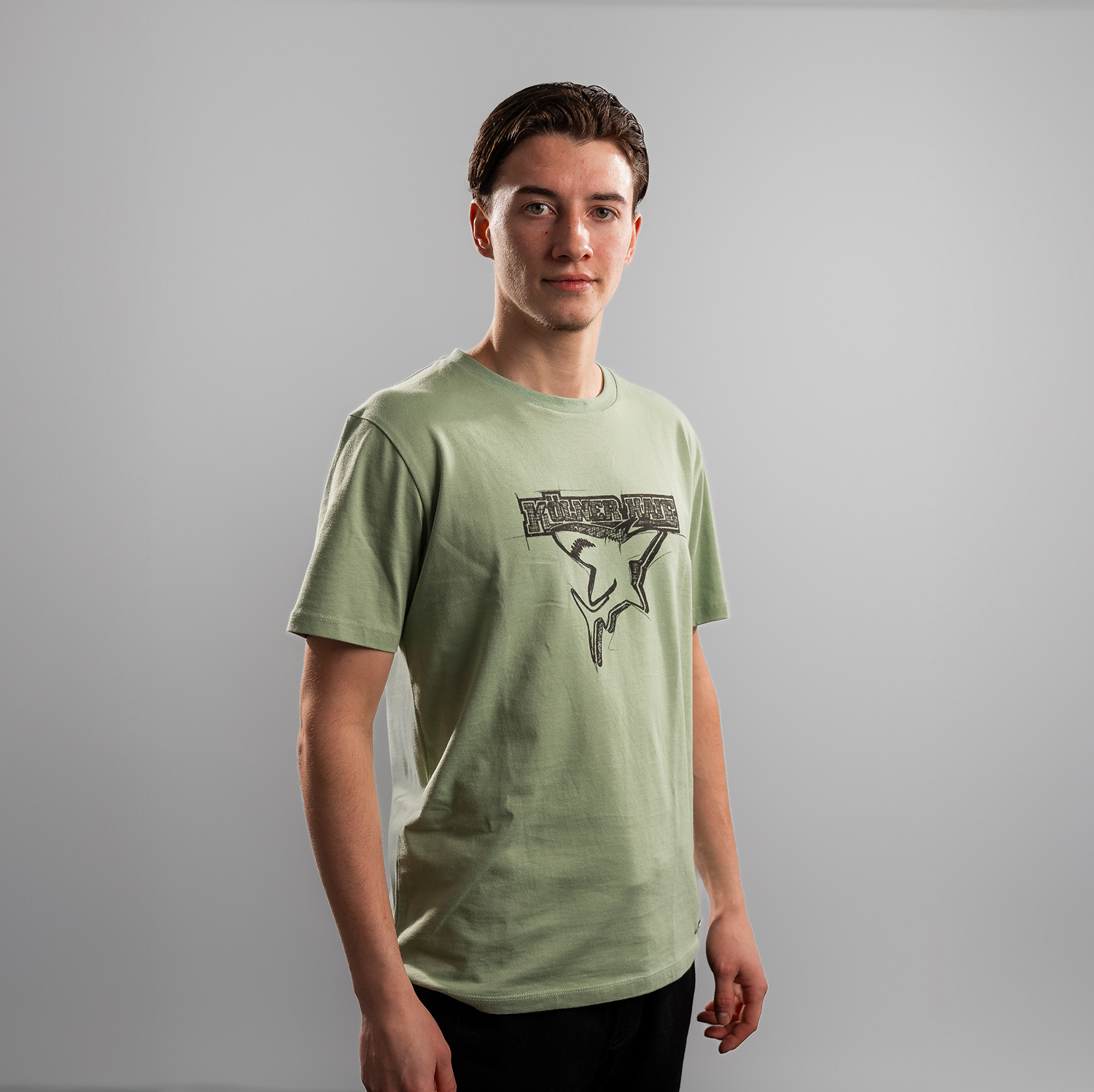 Grünes T-Shirt der Kölner Haie mit großem Frontprint des Kölner Haie-Logos, getragen von Kevin Niedenz, Seitansicht, rechte Schulter vorne, vor hellem Hintergrund.
