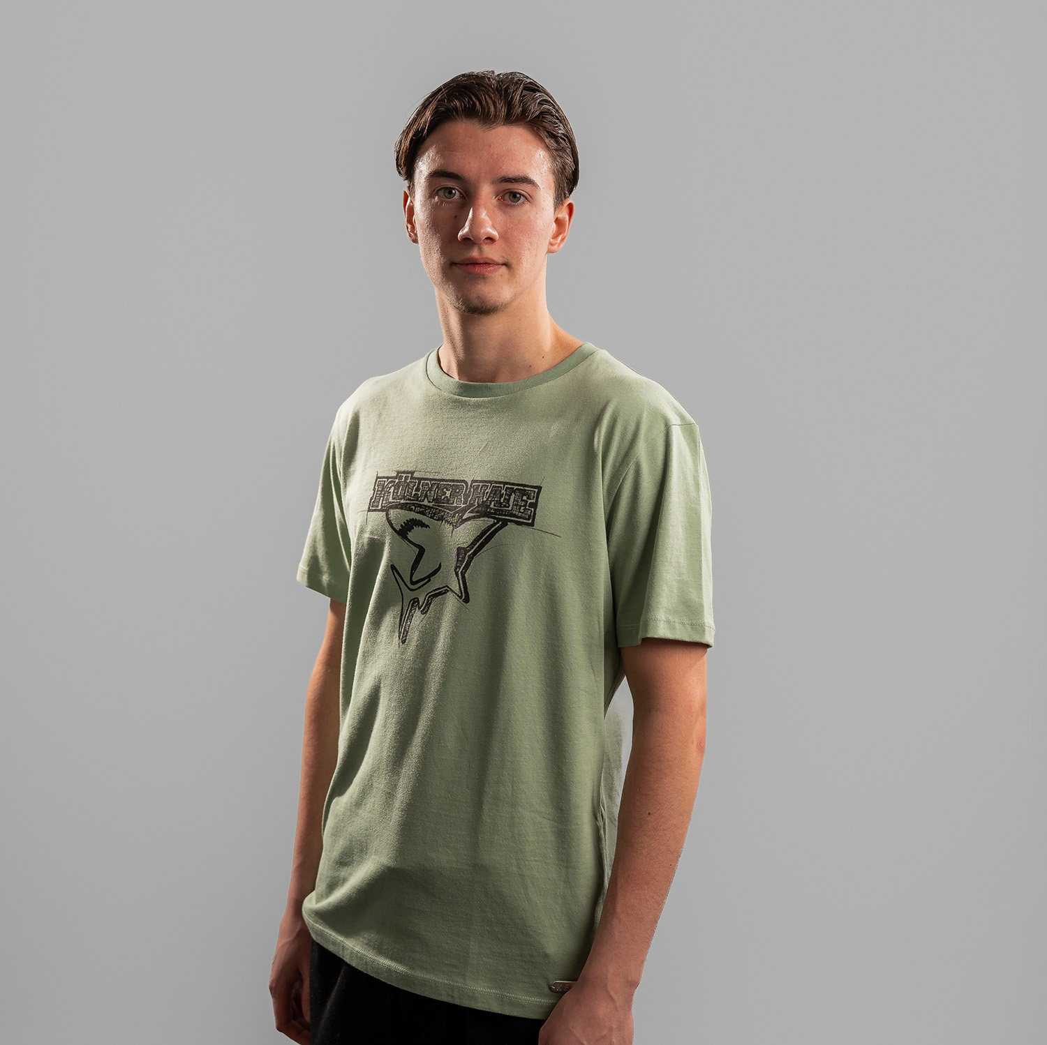 Grünes T-Shirt der Kölner Haie mit großem Frontprint des Kölner Haie-Logos, getragen von Kevin Niedenz, Seitansicht, linke Schulter vorne, vor hellem Hintergrund.