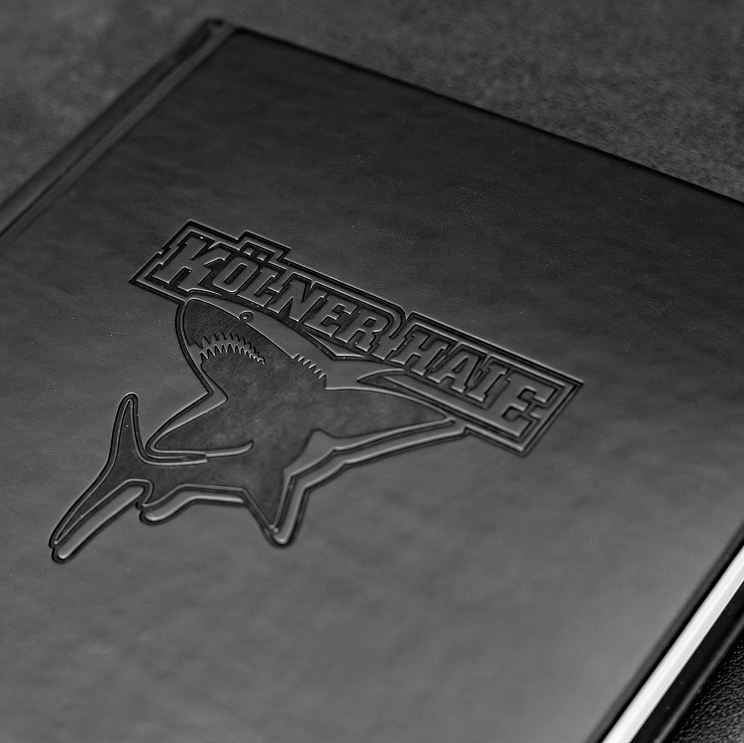 Detailansicht Logo: Schwarzes Notizbuch mit Ton-in-Ton geprägtem Kölner Haie-Logo auf dem Cover