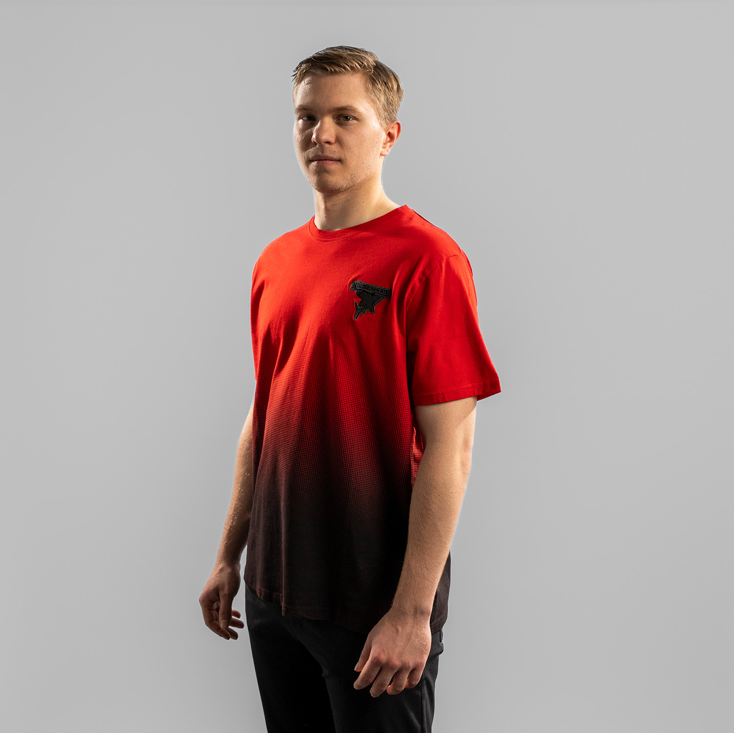 Rotes T-Shirt mit Verlauf ab Brustbereich nach unten zu Schwarz der Kölner Haie mit grau gesticktem Kölner Haie Logo auf der linken Brust (Herzseite), getragen von Tobi Ancicka, Seitansicht. Rechte Schulter vorne, vor hellem Hintergrund.