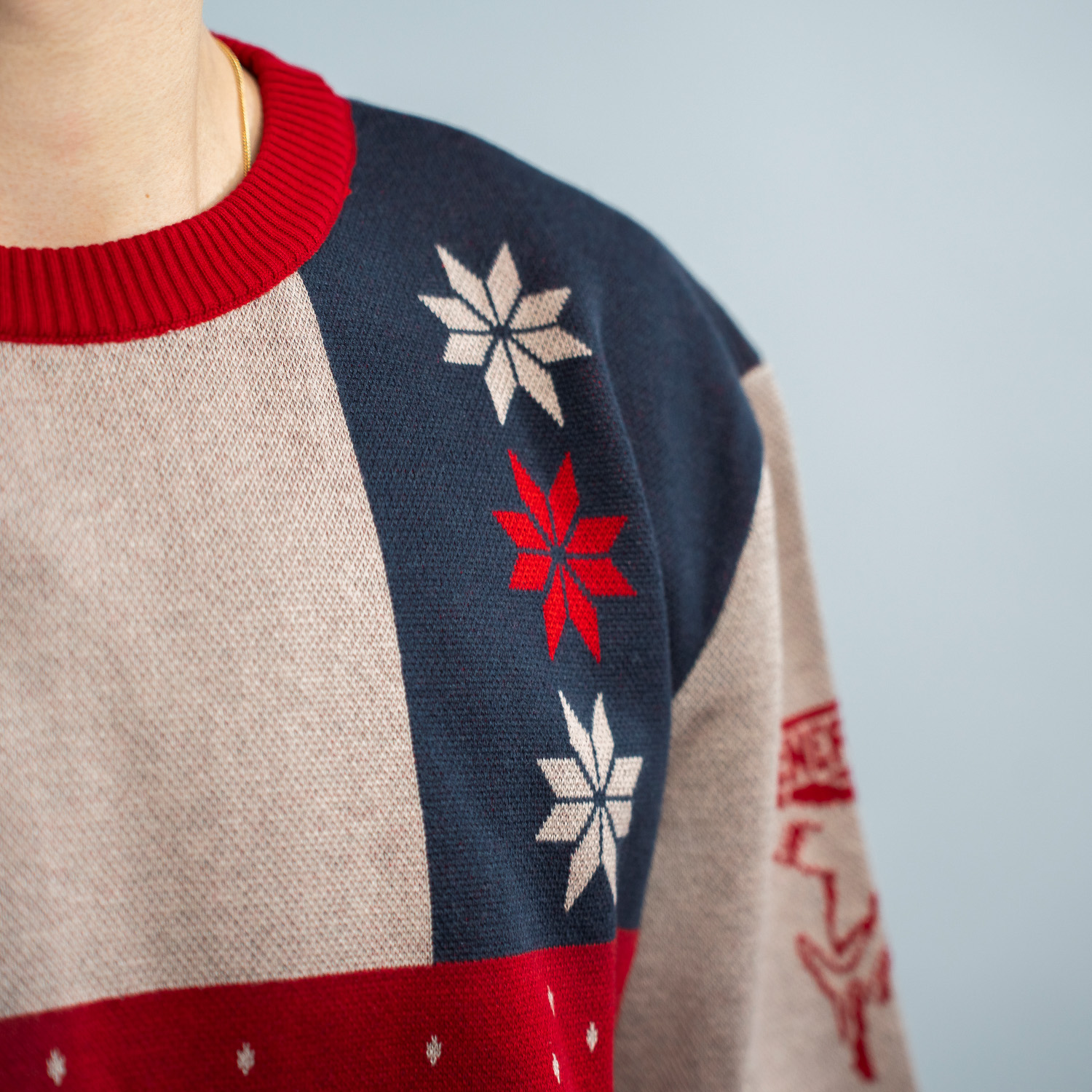 Detailansicht der linken Schultervorderseite des farbenfrohen Weihnachtspullover mit Patchwork-Design in Dunkelblau, Rot, Grün und Beige. Auf einem blauen Part befinden sich untereinander drei achtzackige Nordische Schneesterne, beige-rot-beige. 
