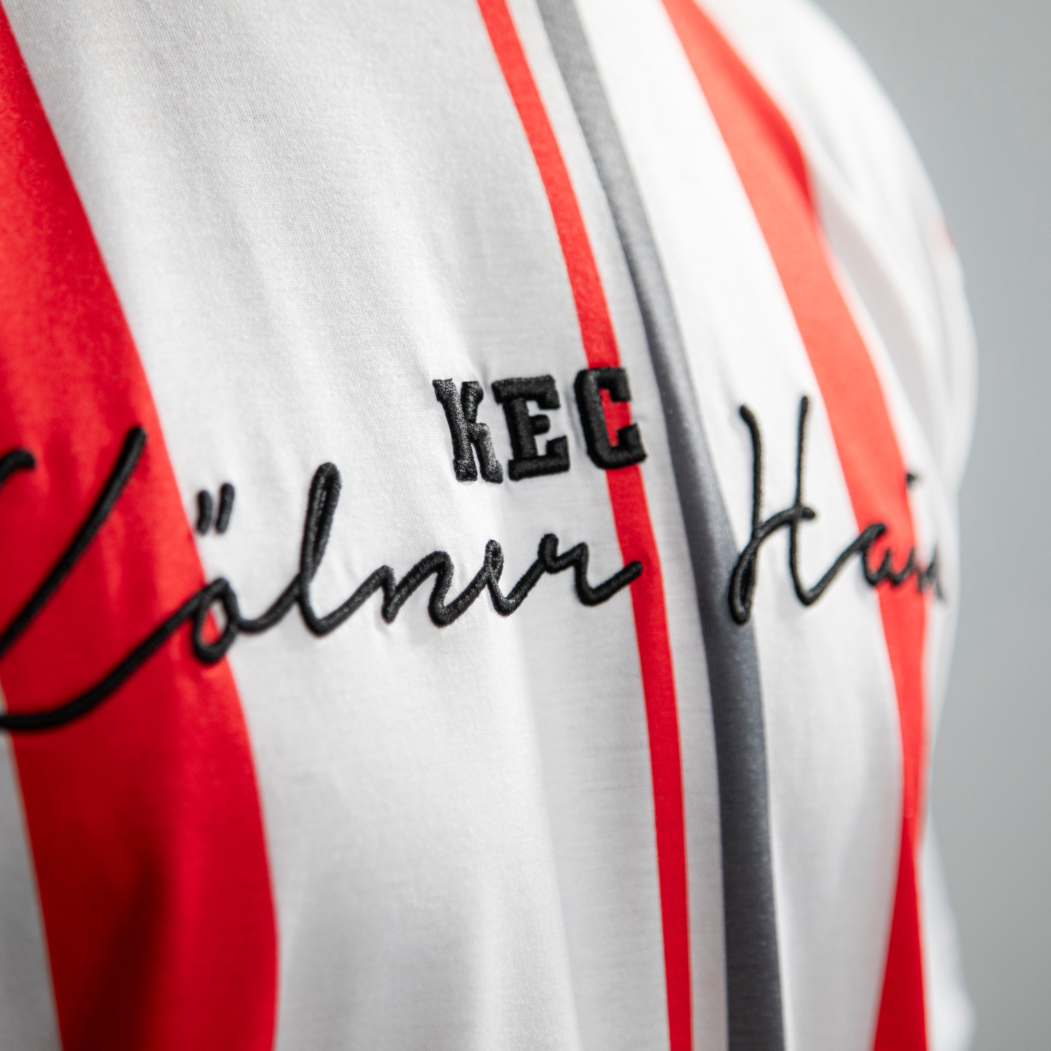 Detailansicht vom schwarzen gestickten Brust-Schriftzug "KEC Kölner Haie" auf dem weißen T-Shirt mit vertikalen Streifen in Rot, Dunkelgrau und Schwarz.