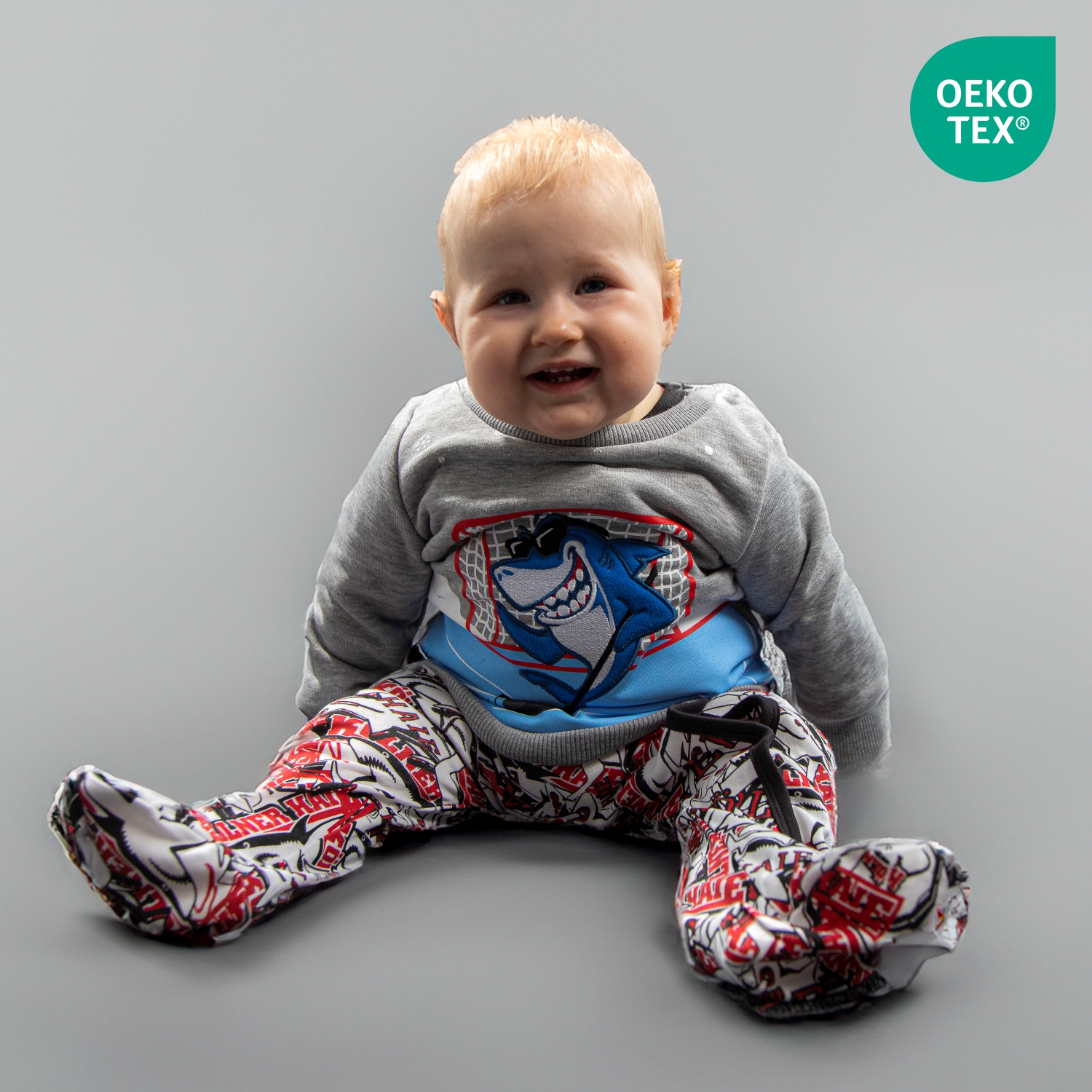 Sweat-Shirt SHARKY Graues Sweatshirt mit großem Frontprint eines lachenden Haies vor einem Eishockeytor, getragen on Babymodel sitzend, vor hellem Hintergrund. Der Artikel ist OEKO-TEX® MADE IN GREEN zertifiziert.