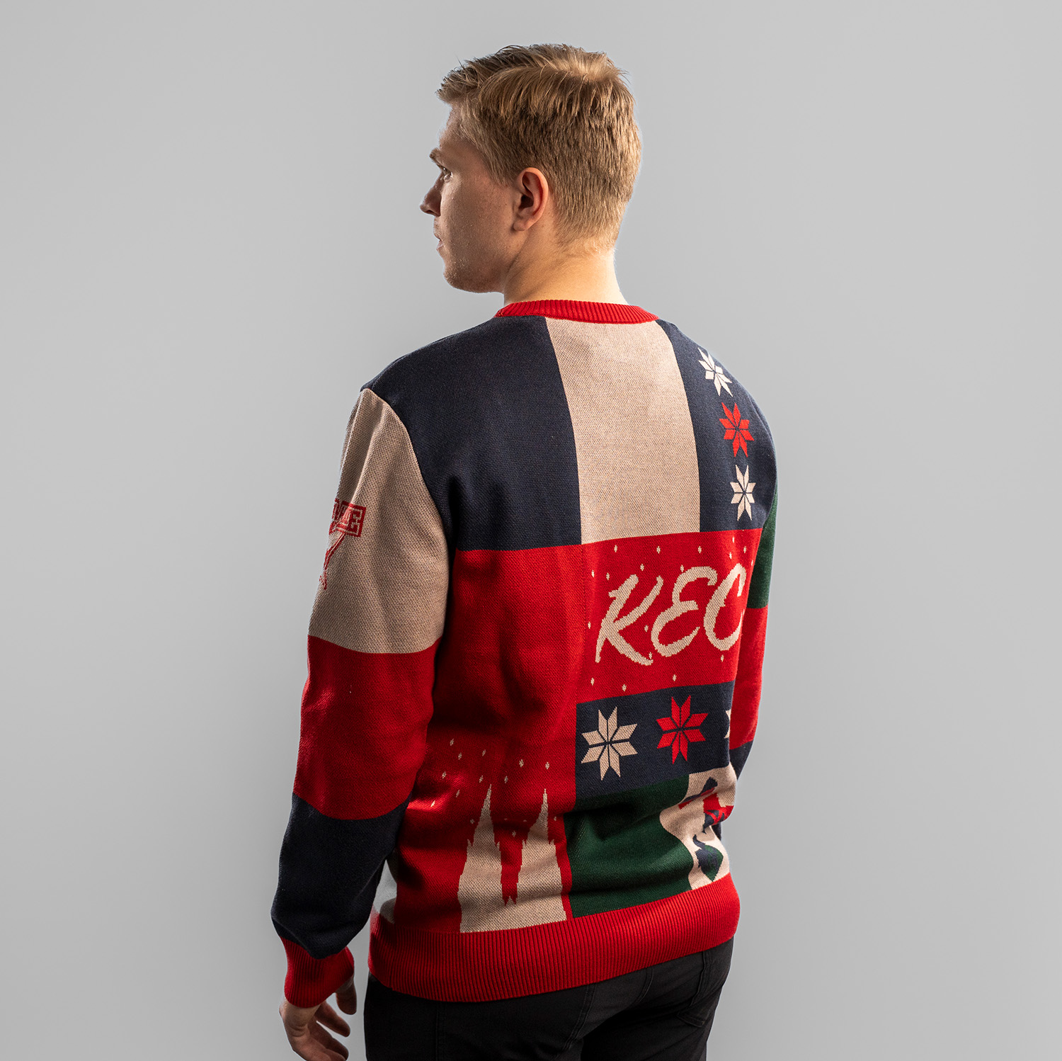 Tobias Ancicka trägt einen farbenfrohen Weihnachtspullover mit Patchwork-Design in Dunkelblau, Rot, Grün und Beige. Auf der Rückseite steht der Schriftzug ‚KEC‘ in geschwungener Schrift, umgeben von Schneeflocken, Geschenken, dem Kölner Dom und stilisiert